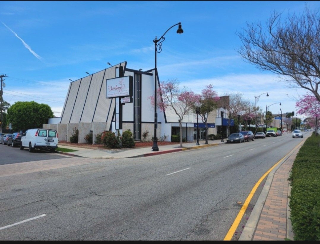 Street-level daytime view of the single-story Destiny funeral home building with its angular white facade and sign, flowering trees along the street