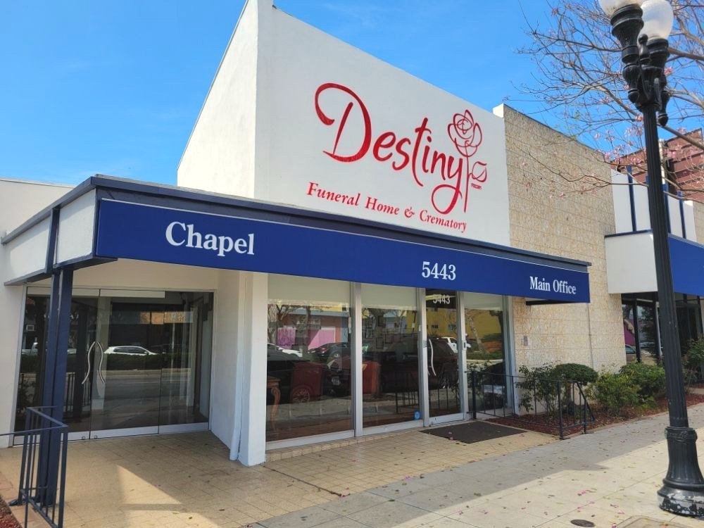 Daytime street view of Destiny Funeral Home & Crematory's white and navy storefront with signage and a flowering pink tree in Long Beach, California.