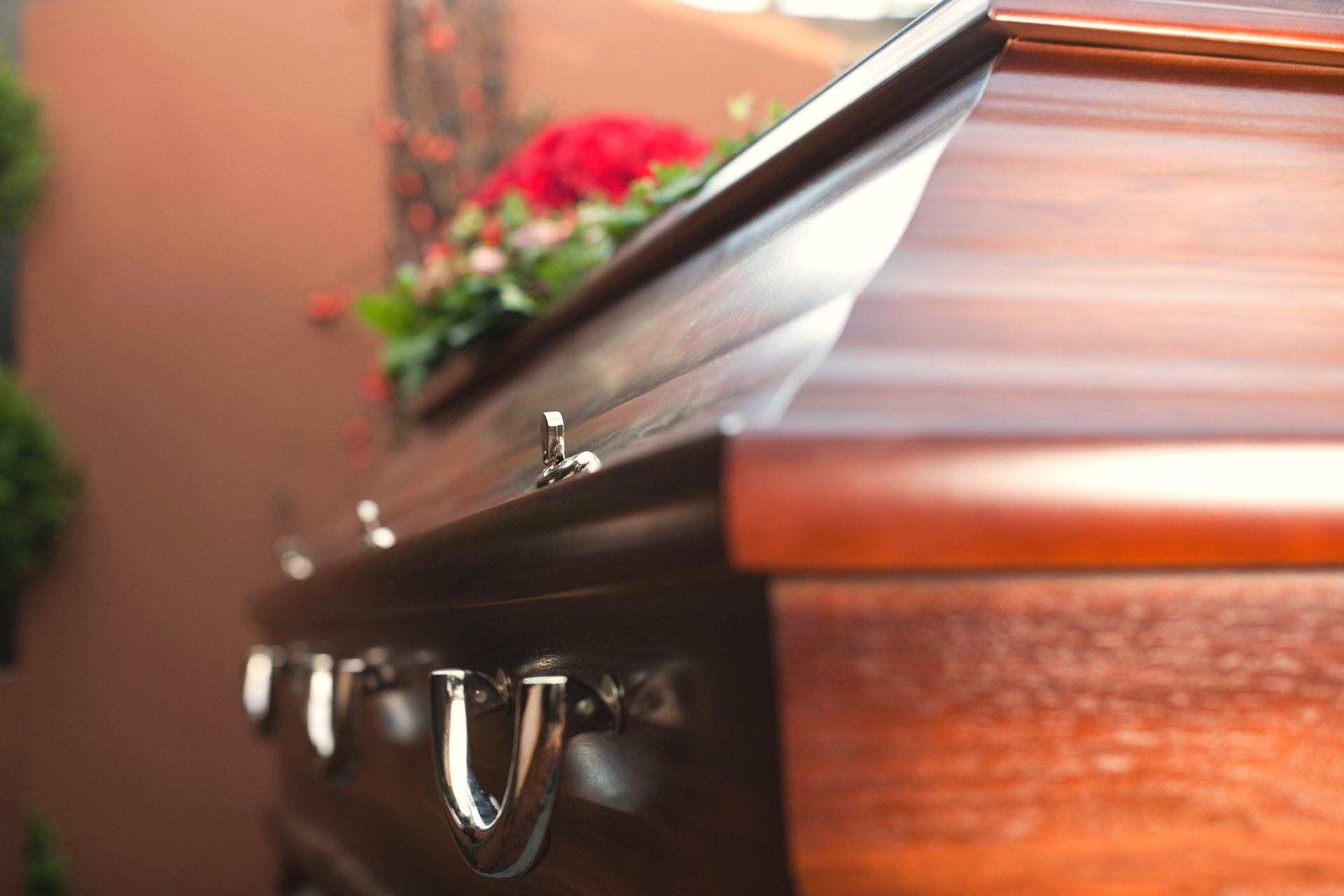 Close-up of a polished dark wood casket with silver handles, softly lit with blurred red and pink flowers in the background.