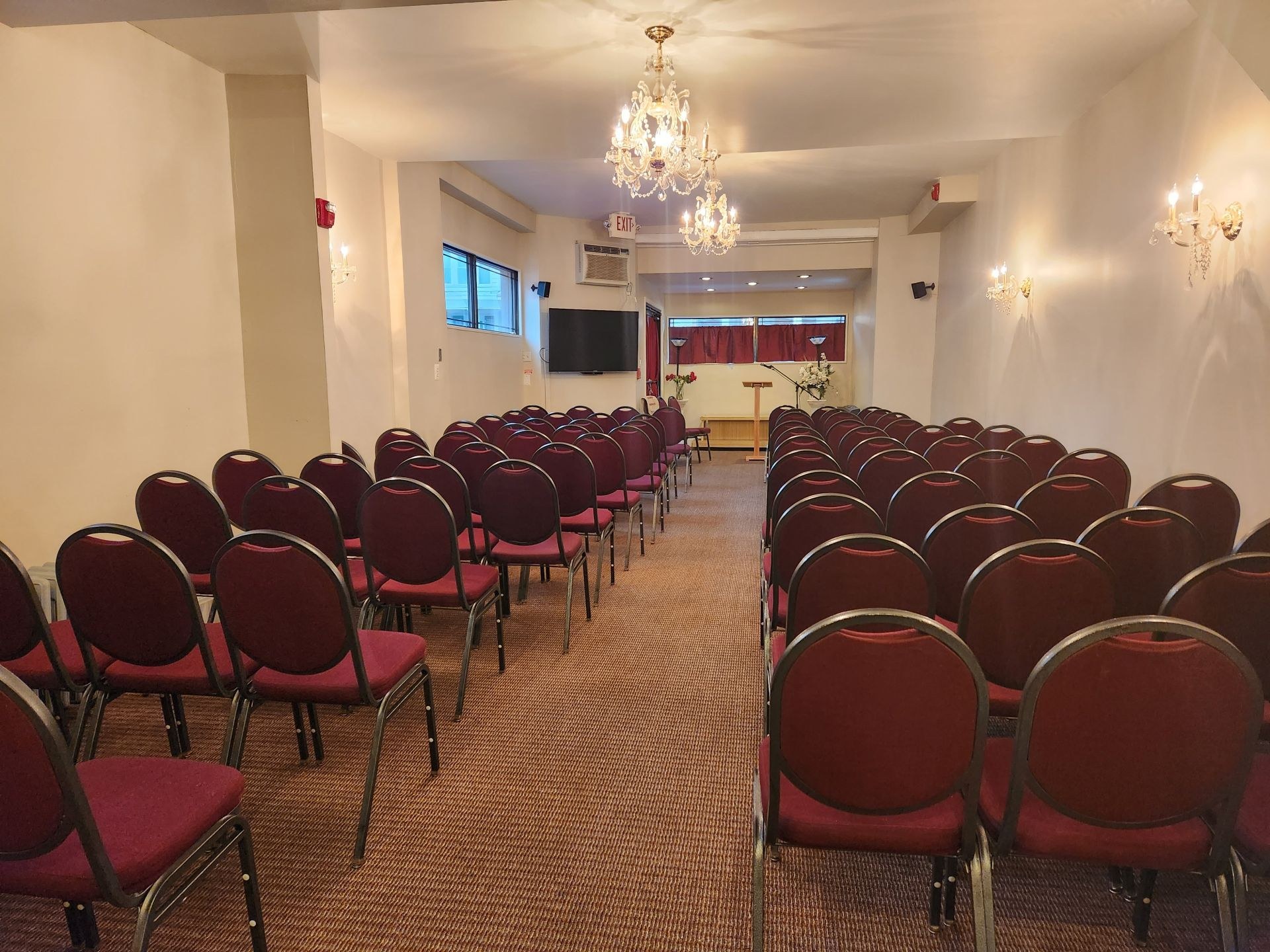 An empty chapel-style room lined with rows of burgundy stacking chairs faces a small podium and floral arrangement beneath ornate crystal chandeliers. Soft indoor lighting and warm wall sconces give the space a formal, hushed readiness before a service begins.
