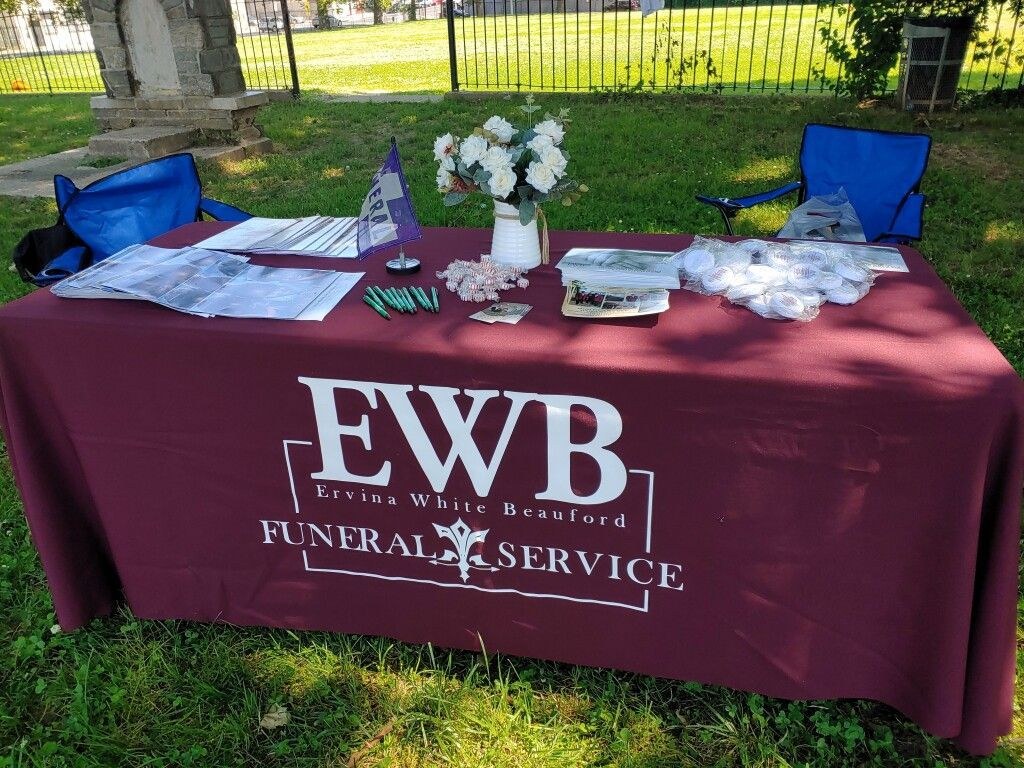 An outdoor community table draped in a maroon cloth printed with a funeral service's name, set with brochures, pens, a white pitcher of faux flowers, and wrapped favors, flanked by empty folding chairs on a sunny grassy lawn near an iron fence.