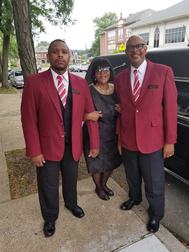 Three funeral staff members in matching maroon blazers stand on a sidewalk beside a black limousine, with a woman in dark dress and sunglasses linking arms between two men, a school building and residential street visible behind them in overcast daylight. The image reads as a staff or service team portrait, formal and composed, with identifiable faces and name badges visible.