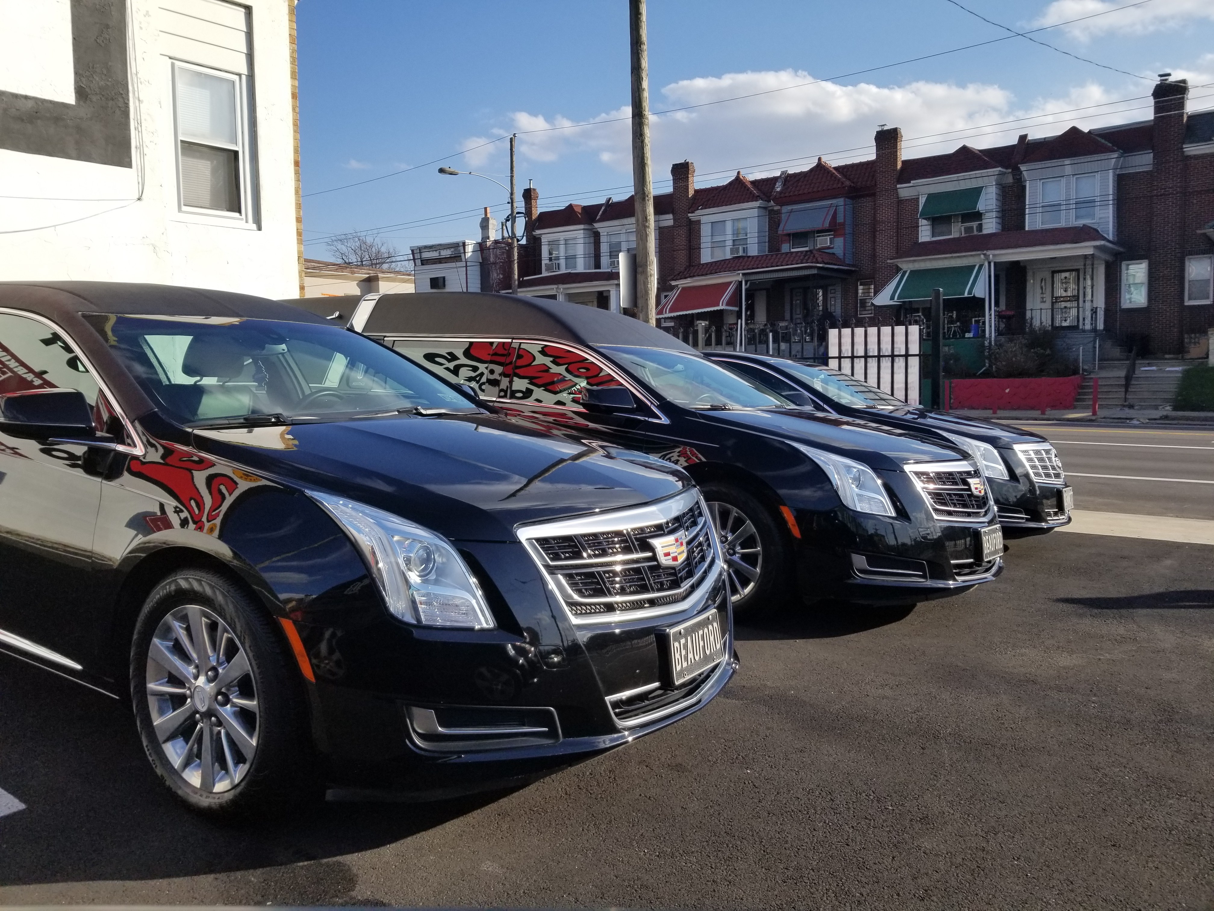 Three black funeral limousines are parked in a row outside a building, with a residential street of brick townhouses and a clear blue sky in the background, in daylight. The image documents a fleet of vehicles used for funeral processions, conveying professionalism and readiness of service.