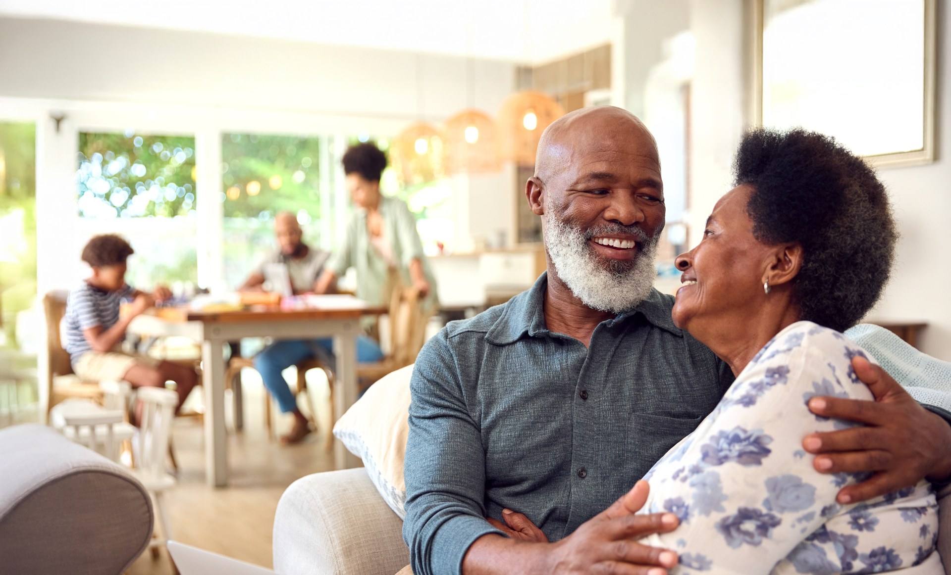 An older man and woman embrace and smile at each other in a bright, modern living room, with a multi-generational family visible relaxing in the background near a dining table.