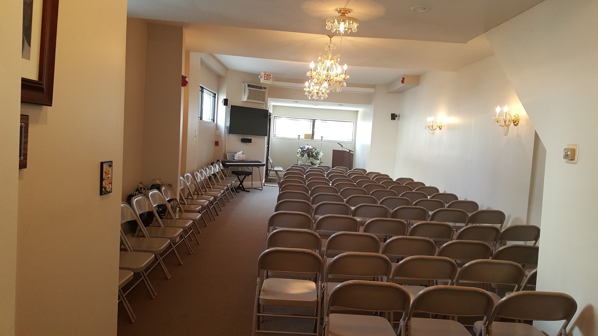 An empty funeral home chapel room with rows of folding chairs facing a podium and floral arrangement, lit by wall sconces and a chandelier, with a wheelchair-accessible ramp and an aisle of extra chairs to the side. The space is arranged and ready but unoccupied, conveying quiet readiness before a service.