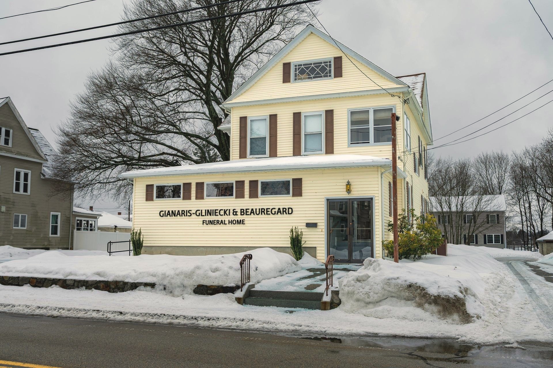 The pale yellow, wood sided funeral home building in Webster, Massachusetts sits on a snow covered corner lot with a shoveled walkway leading to the entrance.