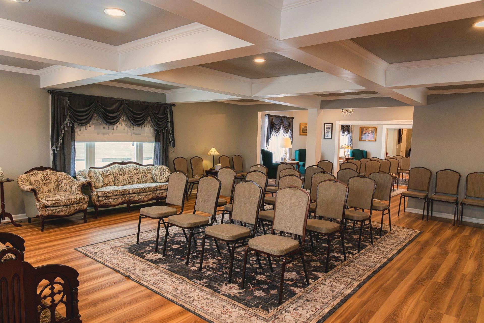 A formal sitting and viewing area at the funeral home with teal wingback chairs, a fireplace, and rows of upholstered chairs arranged for a service.