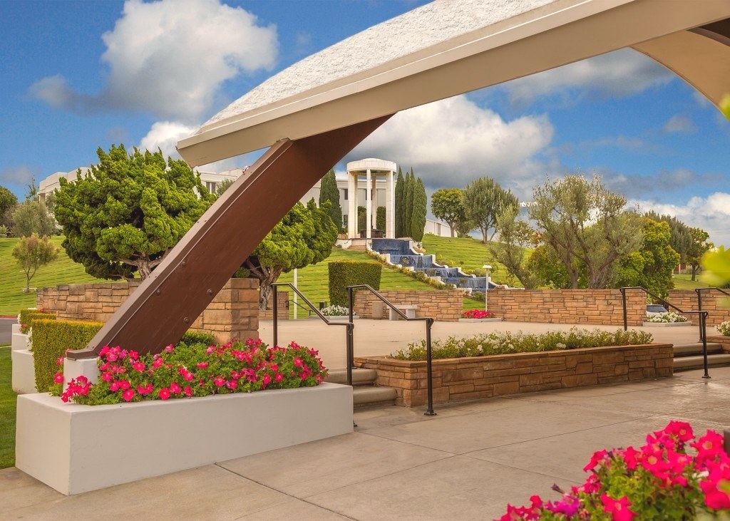 A landscaped memorial park with a modern canopy, stone-columned monument, and cascading water feature beneath a partly cloudy sky.
