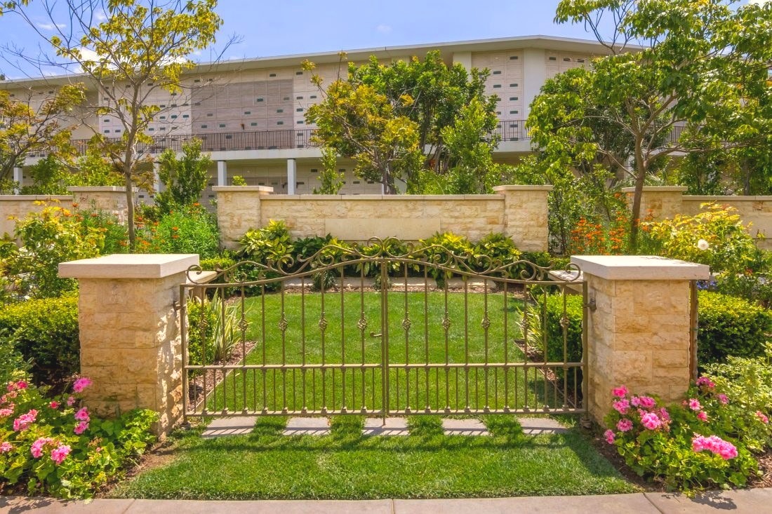 An ornate iron gate stands between stone pillars amid a lush, flowering memorial garden with a tiered mausoleum visible behind it.