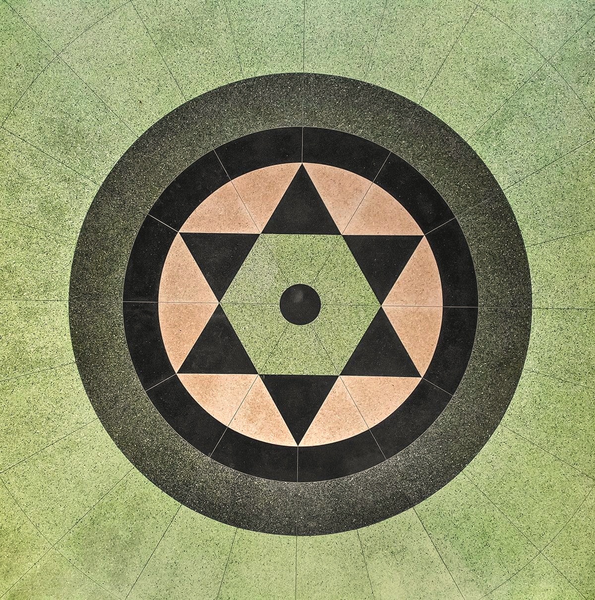 A geometric medallion of a tan and black six-pointed star set within concentric black and green rings on a green terrazzo floor, viewed from directly overhead.