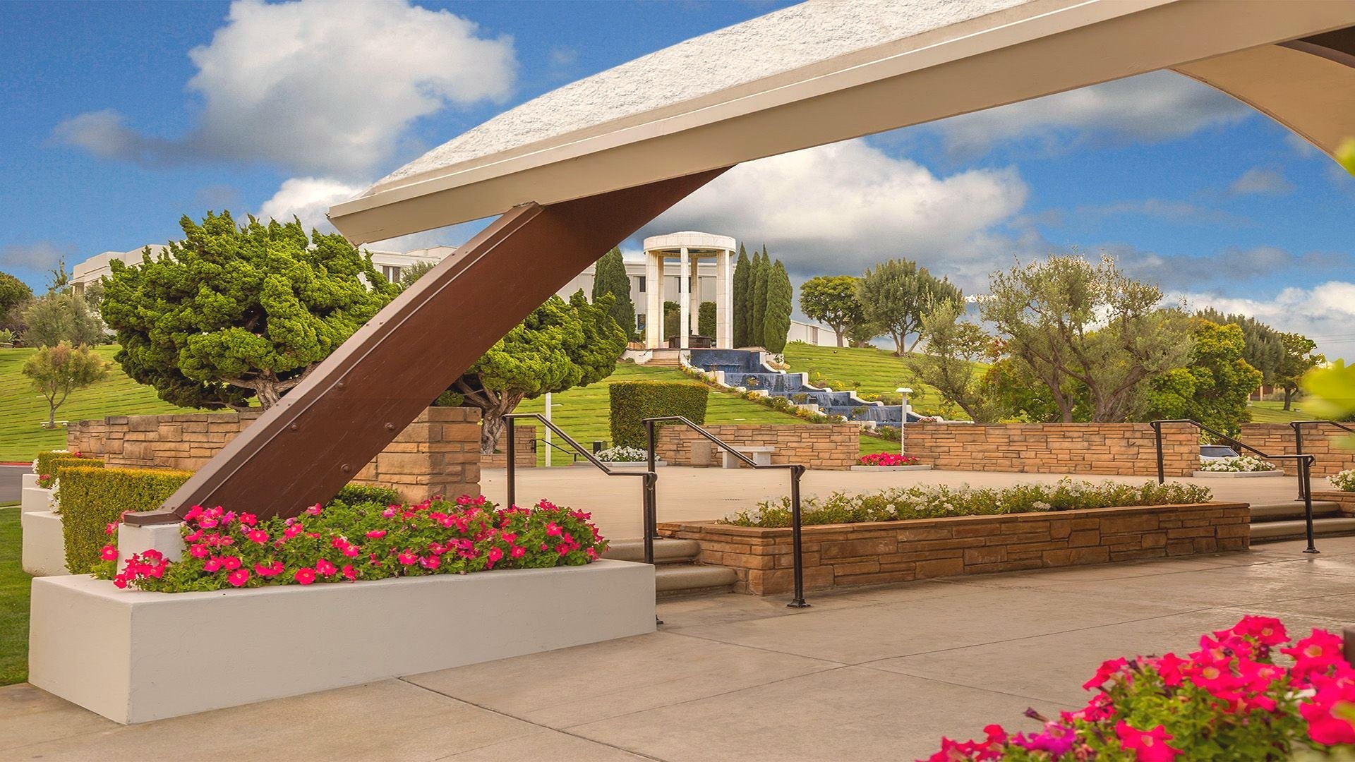 A landscaped memorial park featuring a modern canopy, stone-columned monument, and cascading water feature beneath a bright blue sky with clouds.