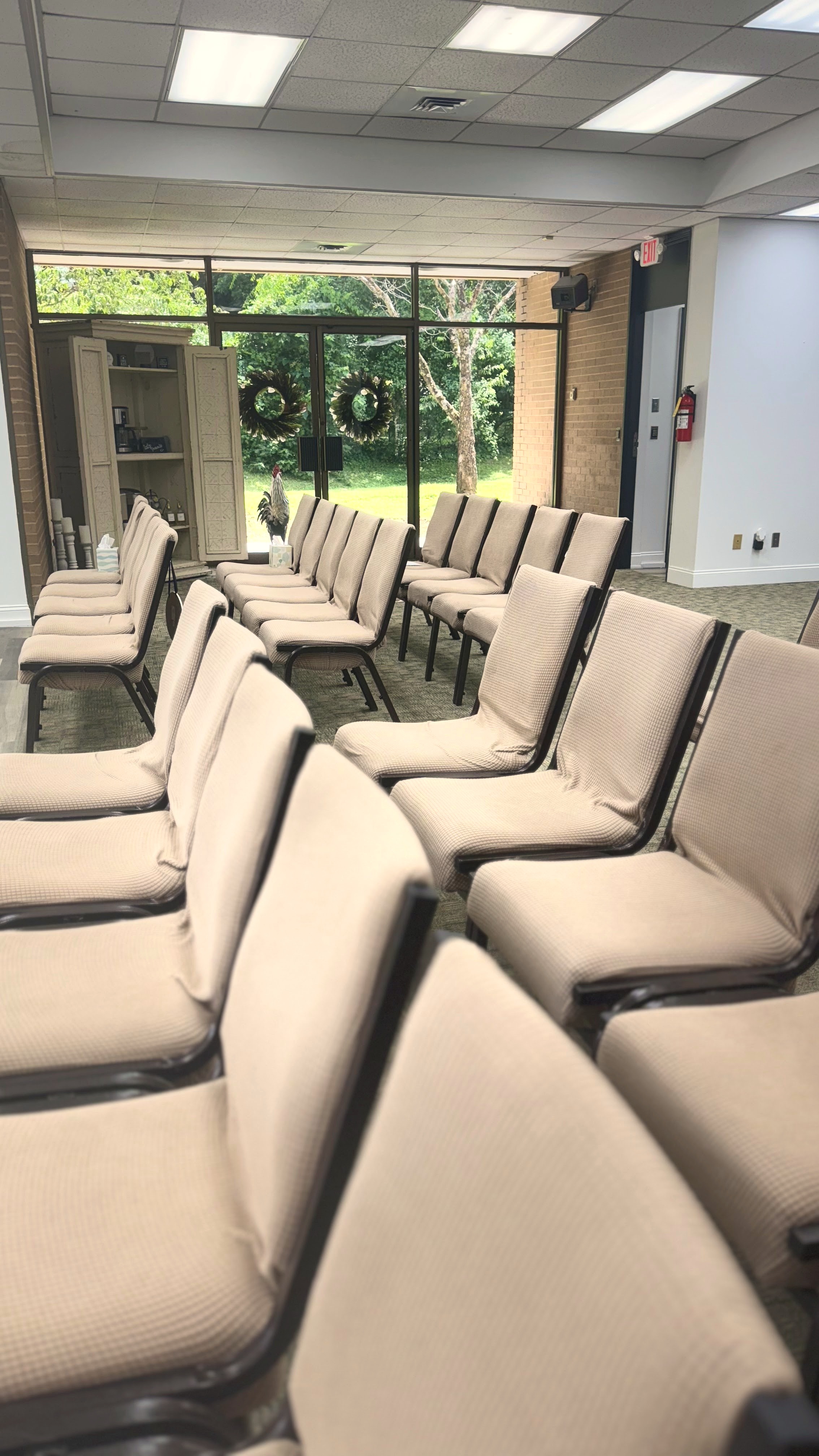 Empty funeral chapel with rows of tan chairs facing glass doors that look out to a green lawn with wreaths