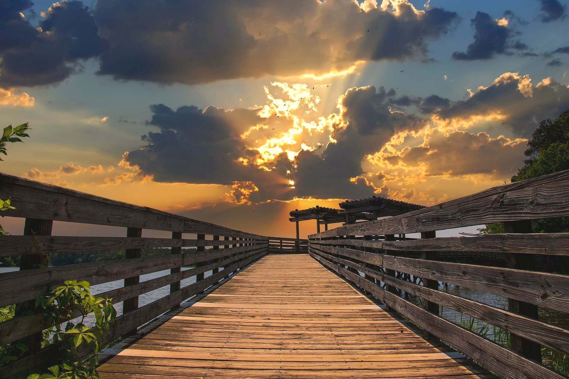 A wooden boardwalk leads toward a pergola over water beneath a sunset sky with sunbeams breaking through heavy clouds.