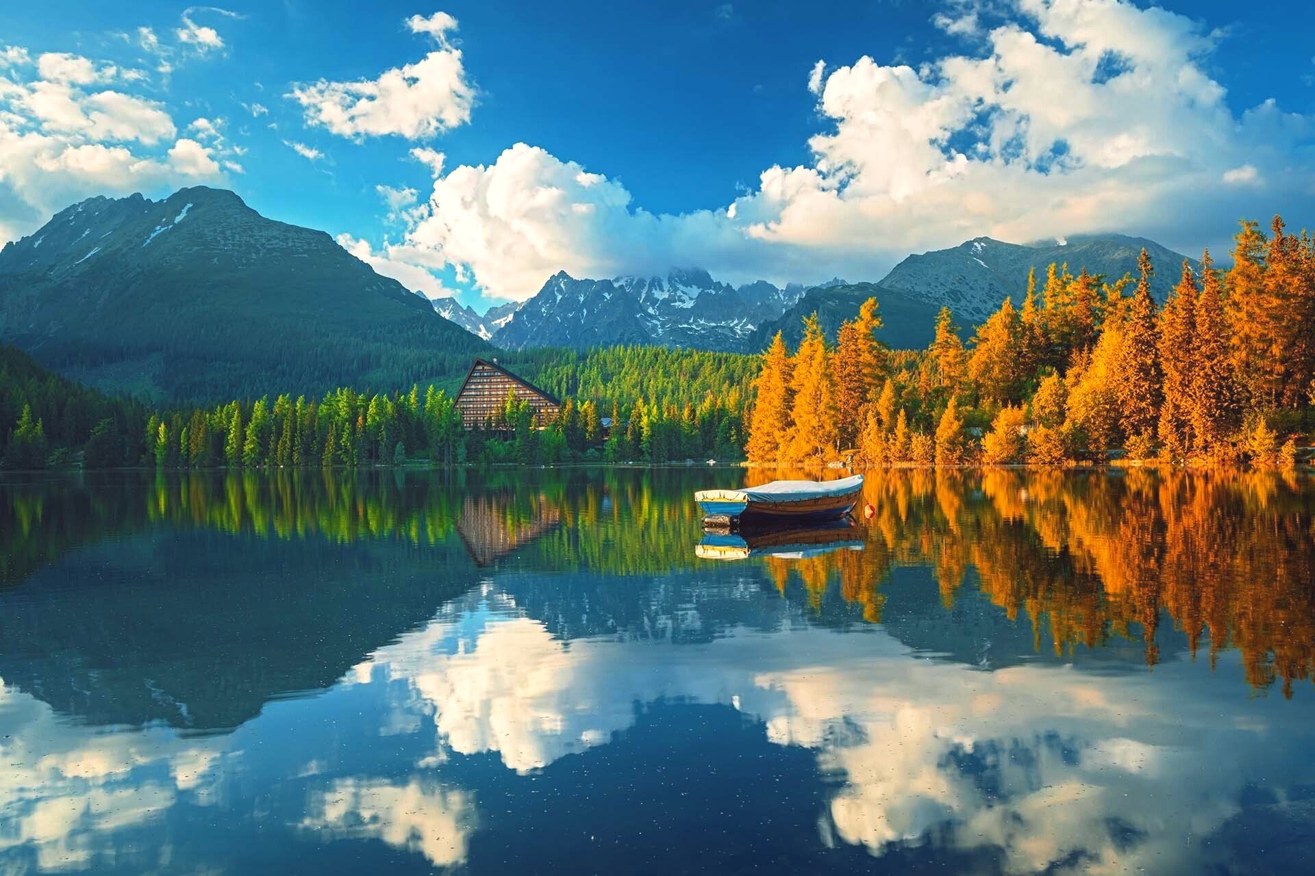 A calm mountain lake reflects a blue sky, snow-capped peaks, and a band of golden autumn larches near a small wooden boat.