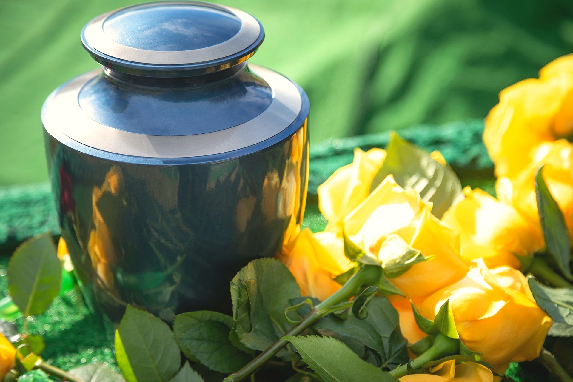 A polished metal cremation urn resting beside yellow roses on a green cloth-draped table, evoking the care Johnson's Funeral Home brings to cremation services.