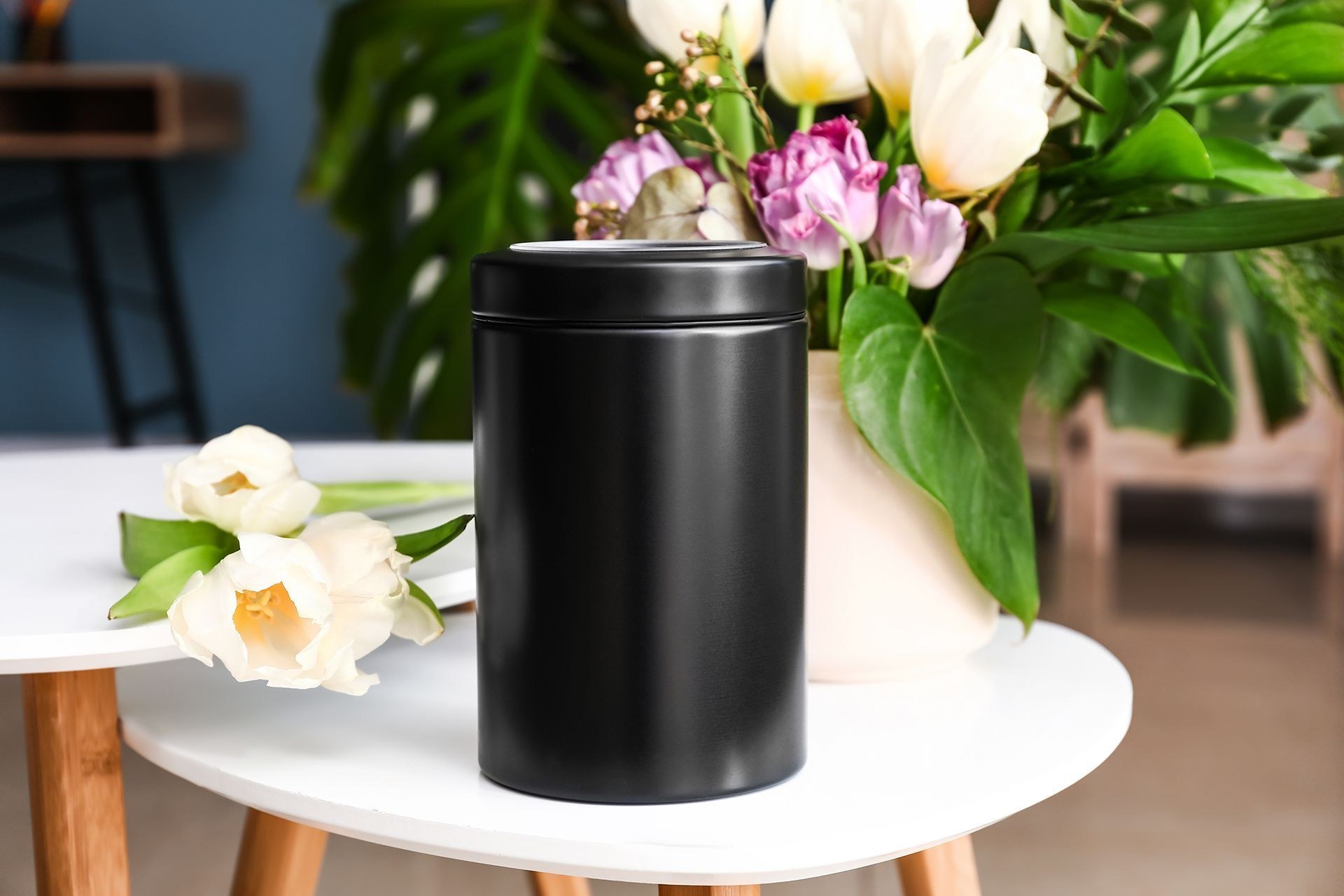 A matte black cremation urn on a round table beside white tulips and pink roses, representing the memorial keepsake and cremated remains options Johnson's Funeral Home offers.