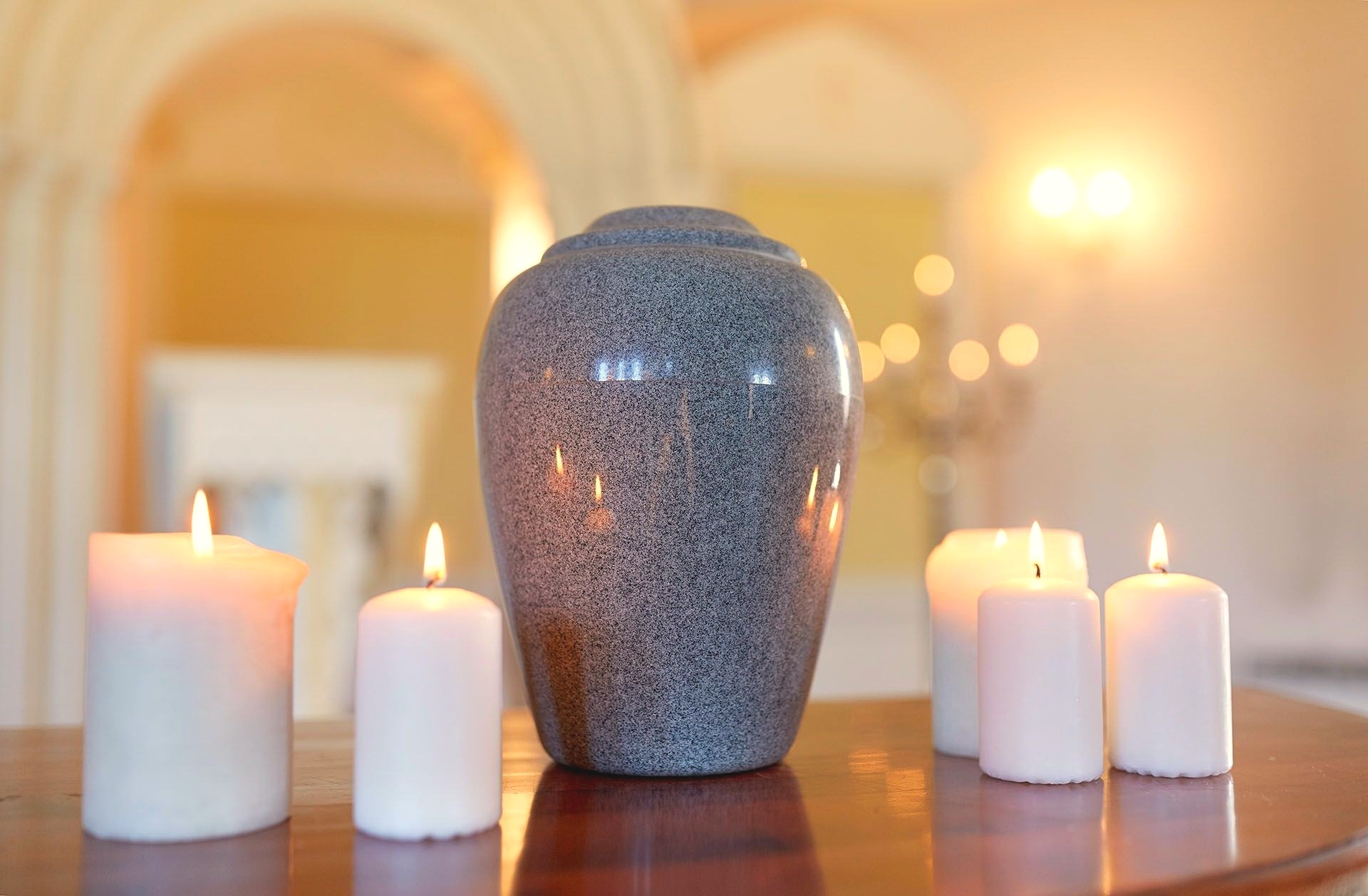 A grey speckled cremation urn rests on a wooden table flanked by lit white pillar candles in a softly lit room.
