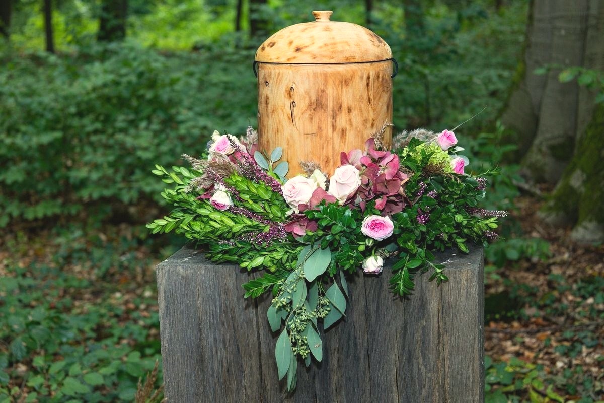 A wooden urn with a domed lid rests on a rustic tree-stump pedestal surrounded by pink roses and hydrangea in a wooded setting.