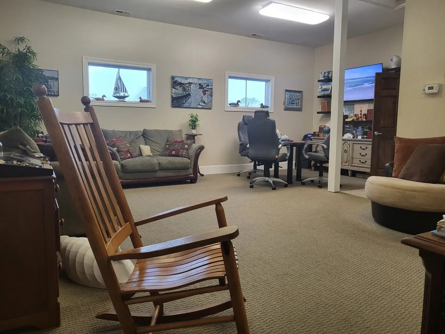 A comfortable lounge area at Noe Funeral Service with a wooden rocking chair, sofas, coastal artwork, and decoy ducks on the windowsill.