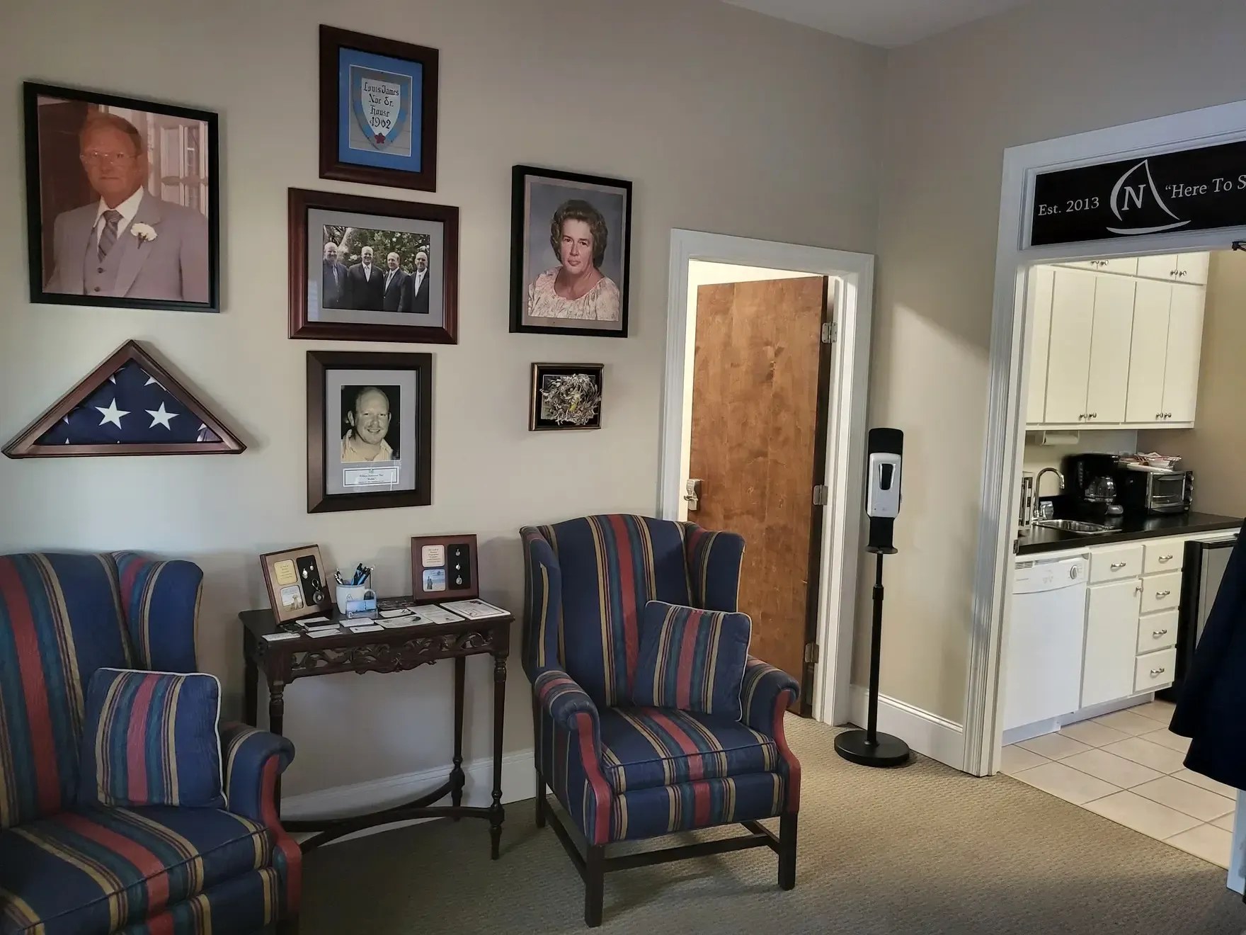 A seating area at Noe Funeral Service with striped armchairs, a wall of framed photographs, a folded flag case, and memorial keepsakes, with a doorway marked 'Est. 2013 Here To Serve.'