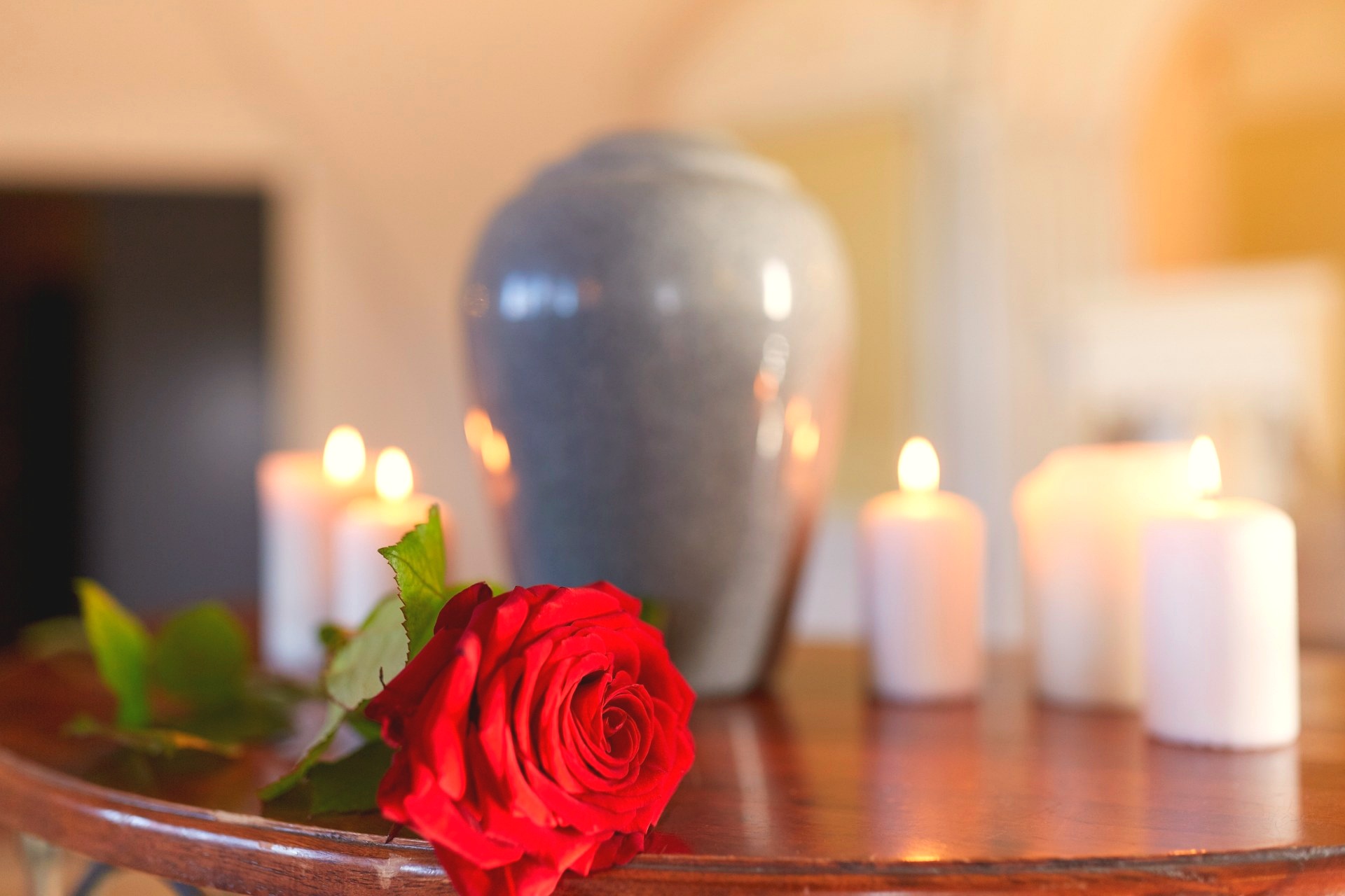 A single red rose lies on a wooden table in front of a grey ceramic cremation urn surrounded by lit white votive candles.