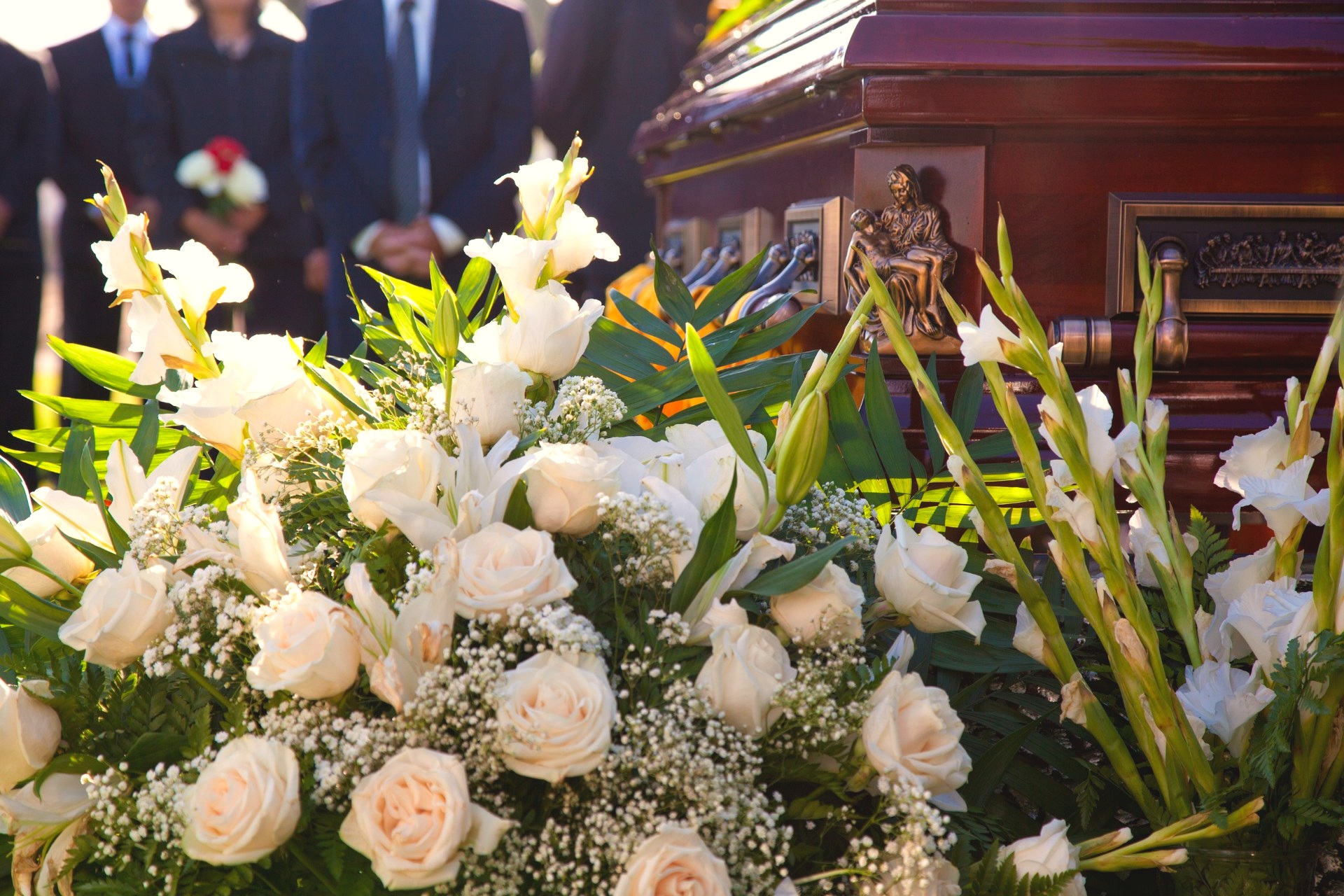 White roses, lilies, and gladioli are arranged beside a dark wood casket with mourners standing blurred in the background at an outdoor service.