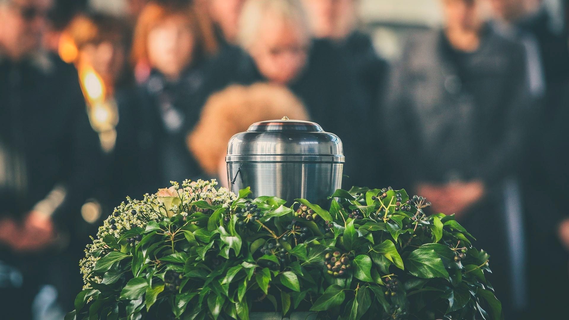 A polished metal cremation urn rests on a wreath of dark green ivy leaves and berries, with mourners gathered softly out of focus in the background.
