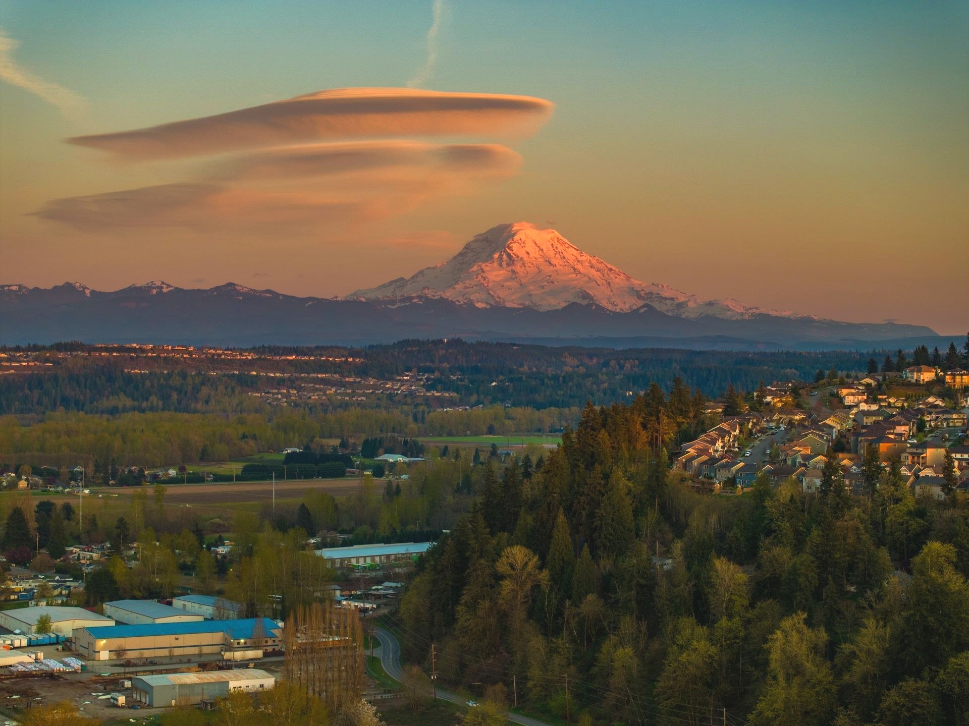 Aerial dusk view of a snow capped mountain glowing pink above a valley town with scattered buildings and forested hills.