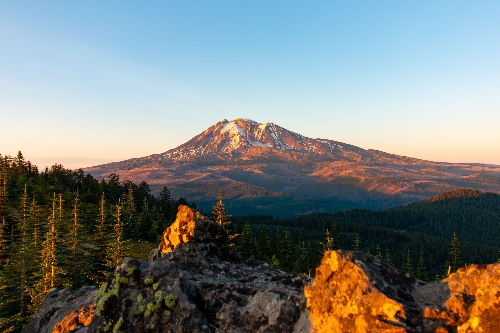 Snow covered volcanic peak glowing orange and gold at sunset above dark forested ridgelines.