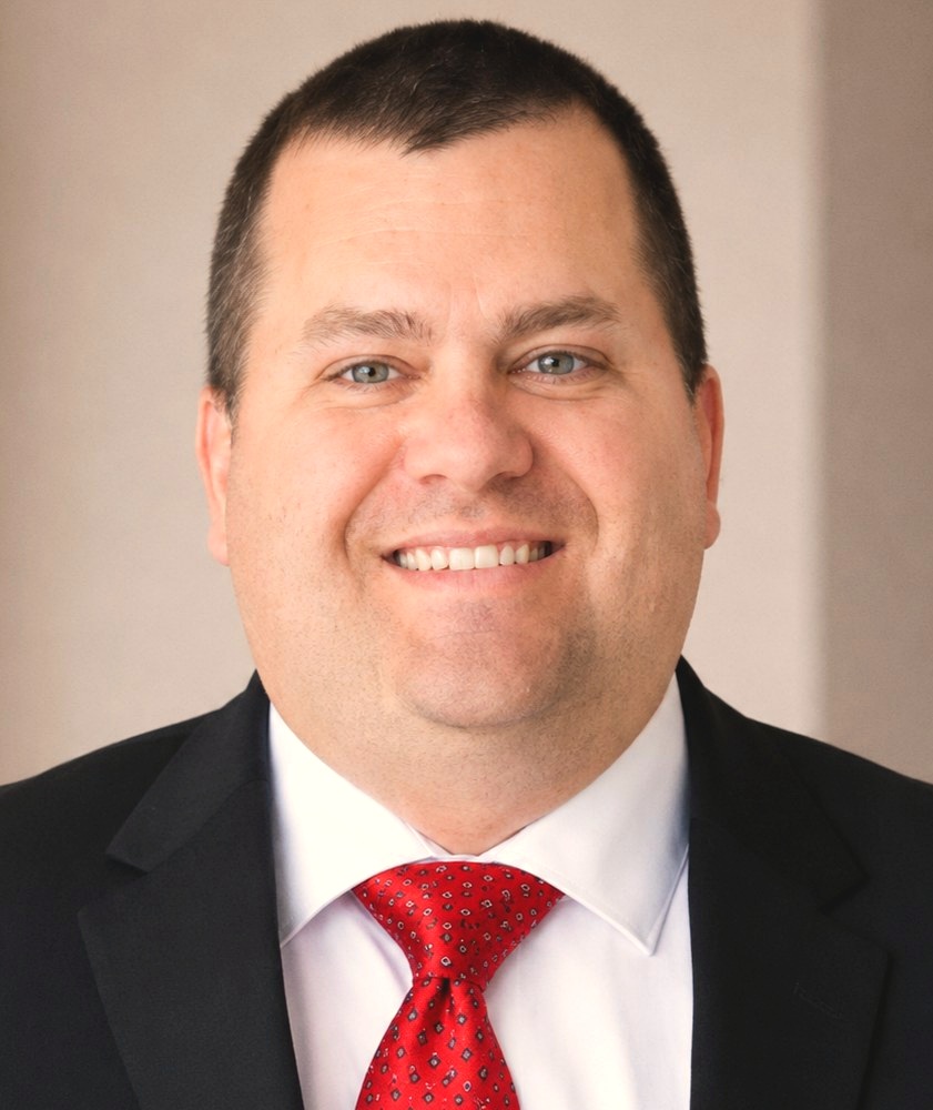 A smiling man with short dark hair wearing a dark suit and red patterned tie, photographed against a plain light-colored studio backdrop, representing a Walker Sanderson funeral director.