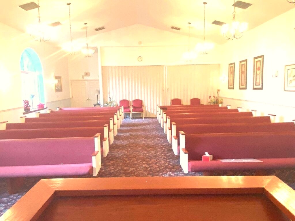 An empty chapel interior with rows of maroon pews facing a curtained alcove, lit warmly by chandeliers and wall sconces, with a wooden podium in the foreground.