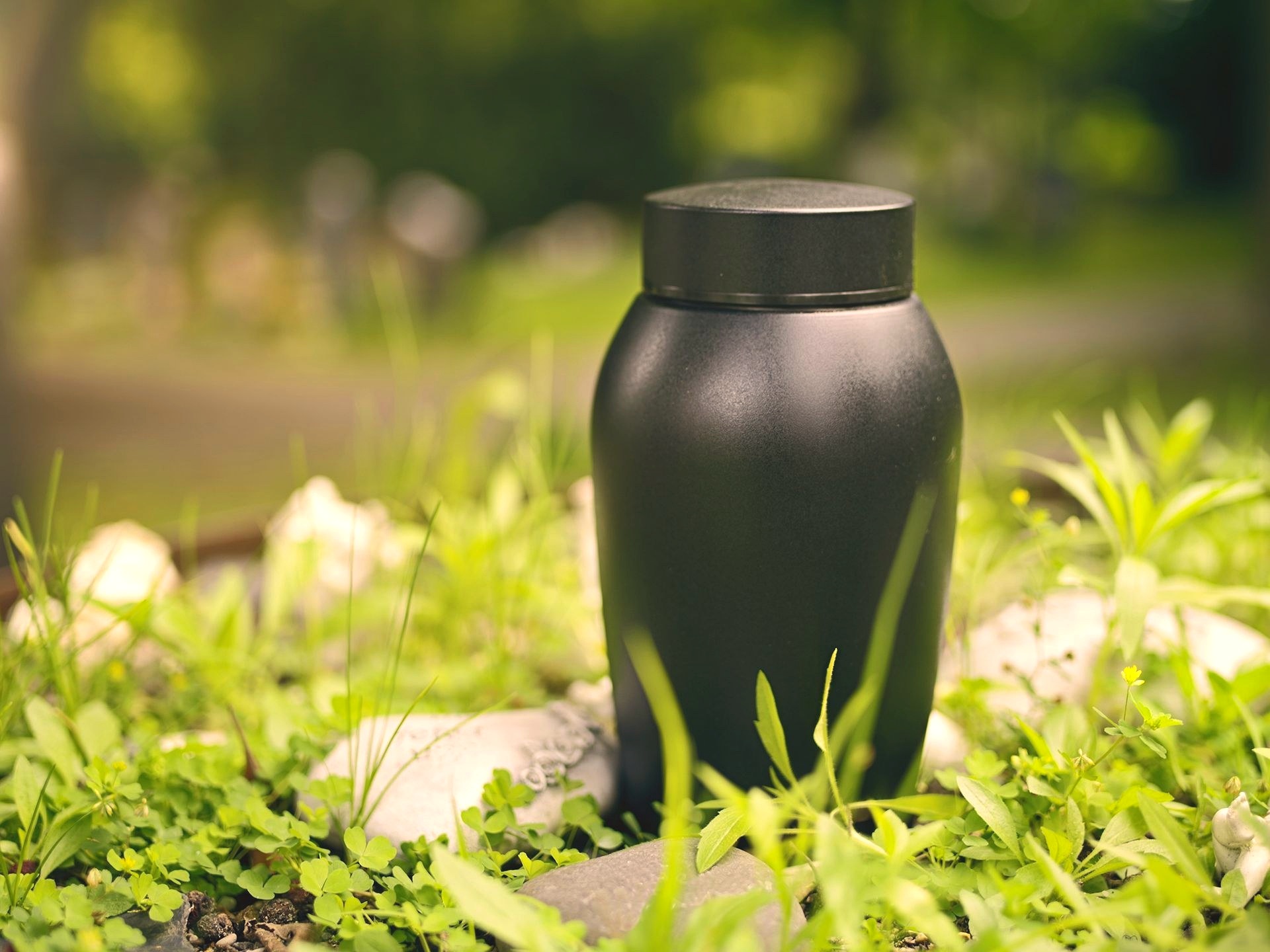 A small dark memorial urn resting outdoors on stones and greenery in a softly lit garden setting.