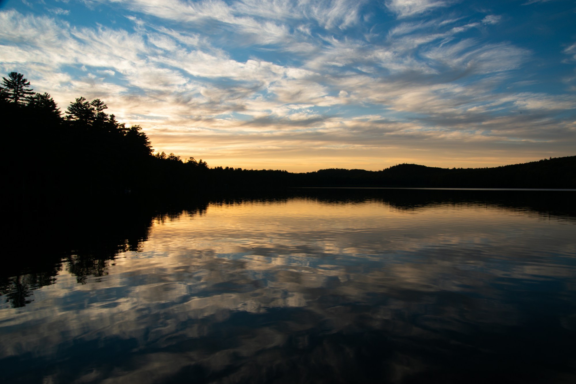 A tranquil lake reflects a colorful dusk sky, surrounded by dark tree silhouettes.