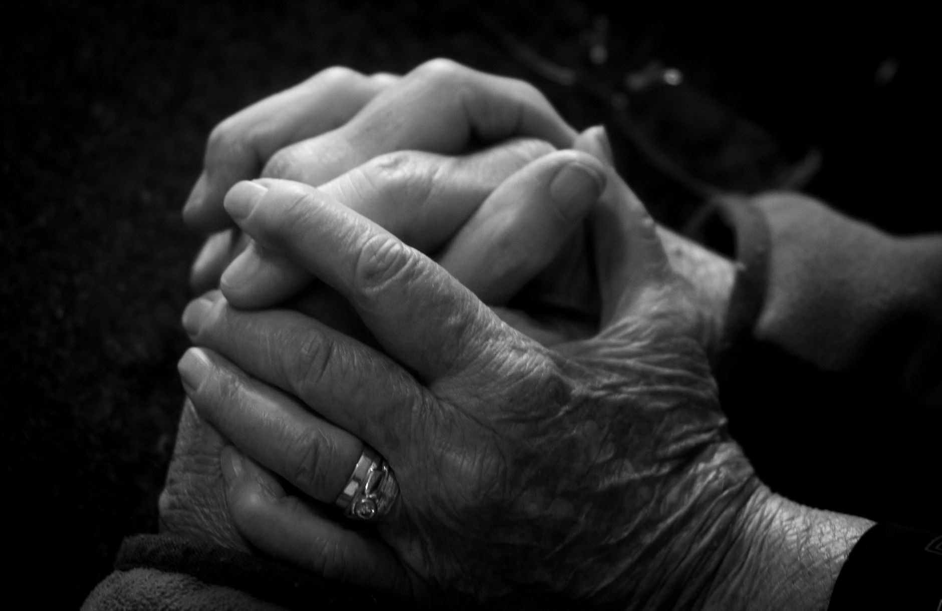 A close-up black and white photograph of two elderly hands clasped together, one wearing a ring, conveying gentle connection and remembrance.