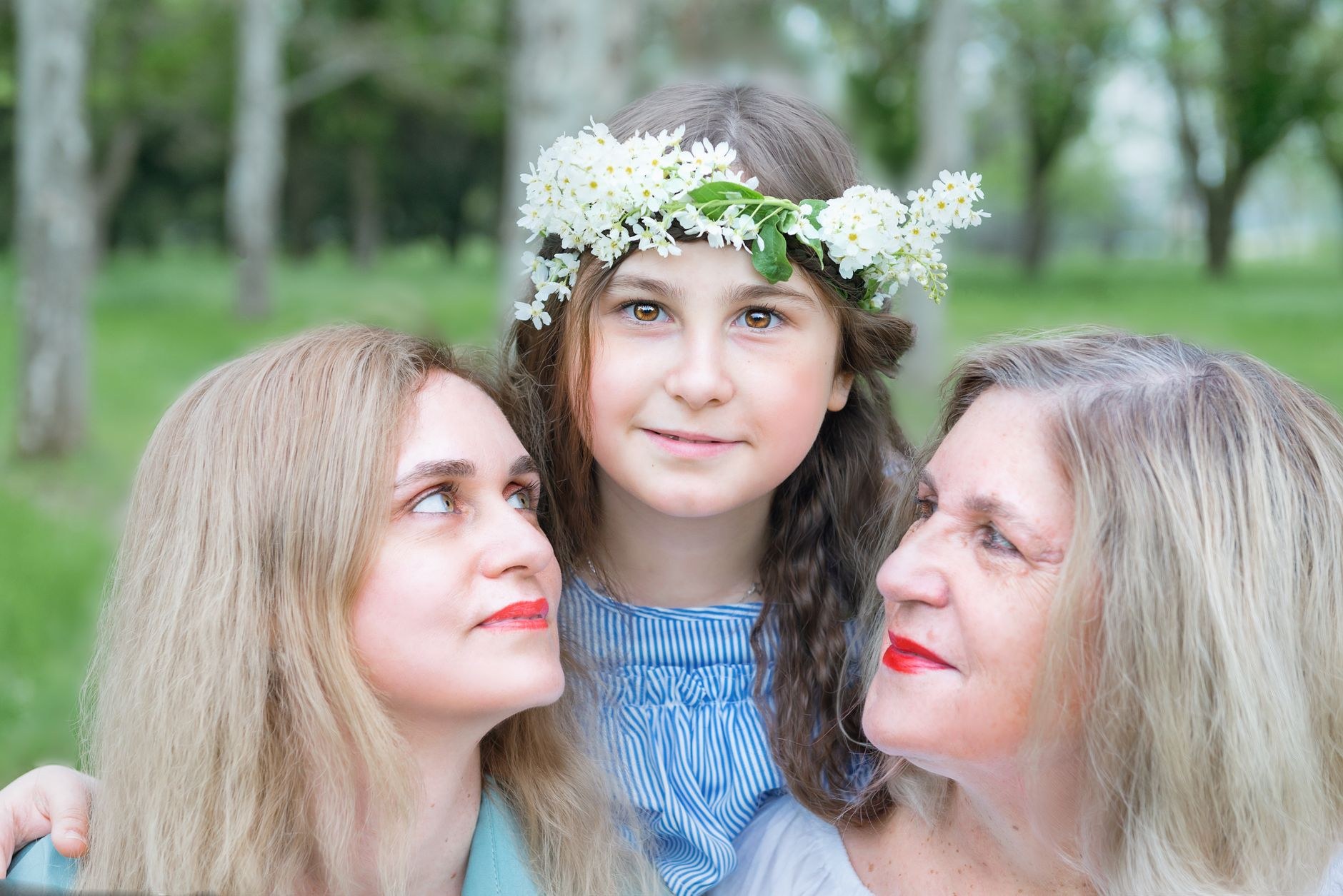A young girl wearing a floral crown stands between two women, one younger and one older, both looking up at her with affectionate expressions in a green outdoor setting, likely spring or summer. The composition suggests three generations of a family and conveys tenderness, care, and generational connection.