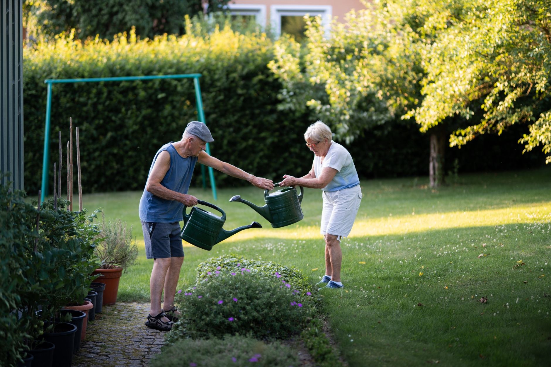 An older couple waters plants together in a backyard garden, suggesting care, continuity, and the peaceful memories families hold.