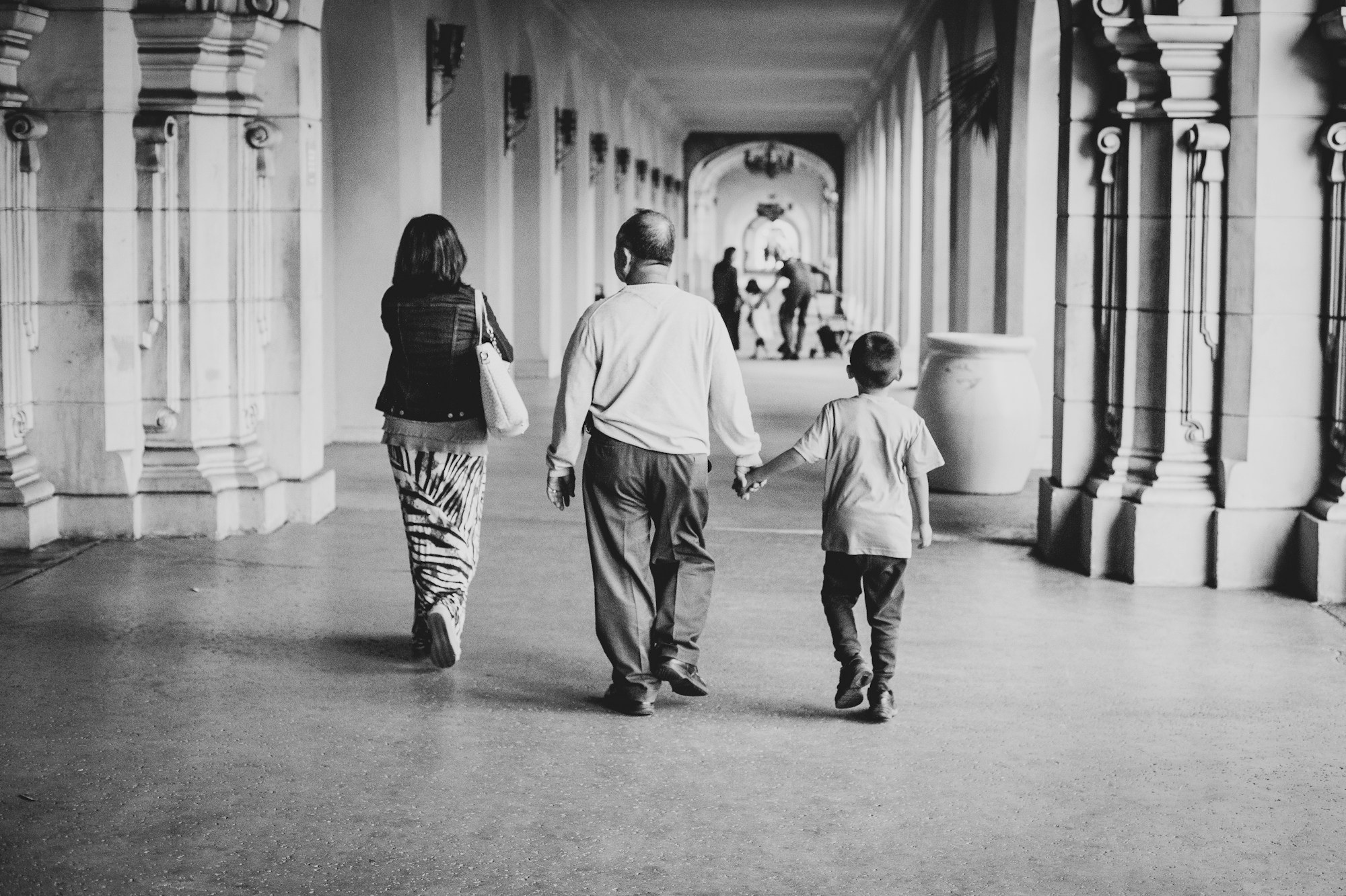 A black-and-white photo of an older man holding a young boy's hand as they walk together through an outdoor colonnaded corridor, representing family and generations across Utah County.