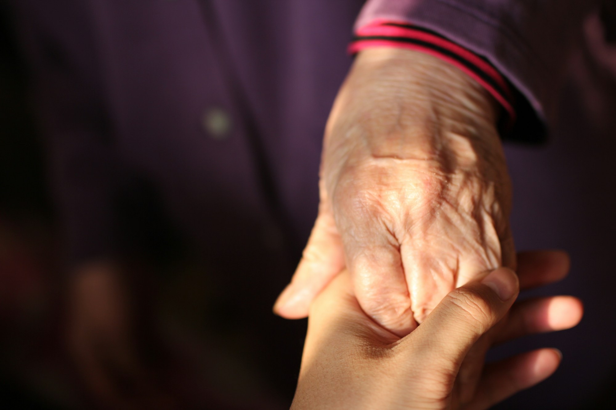 A child's hand holding an elderly person's hand against a blurred reddish-brown background.