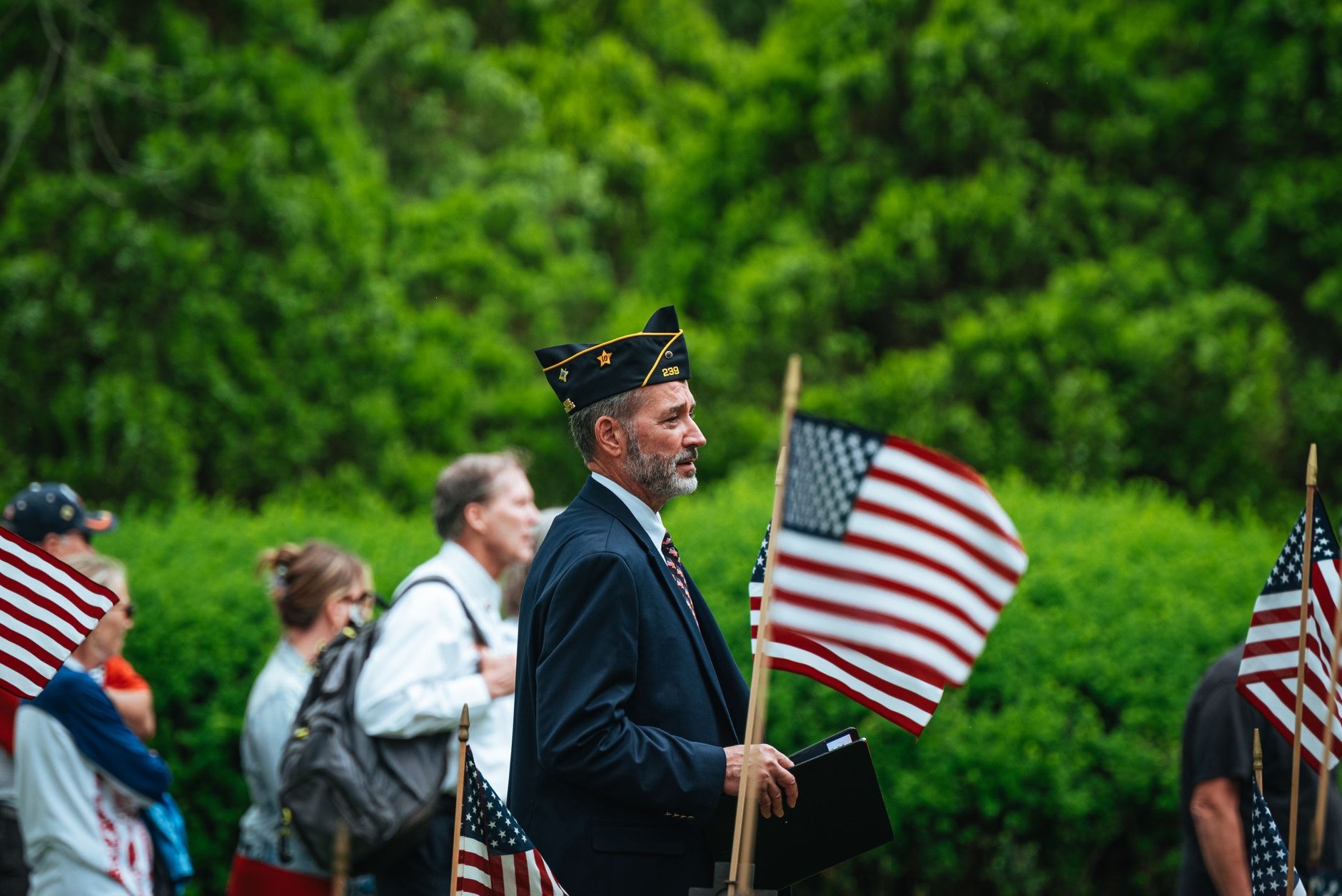 A man in a veteran's cap and white uniform shirt is seen from behind, saluting during an outdoor ceremony.