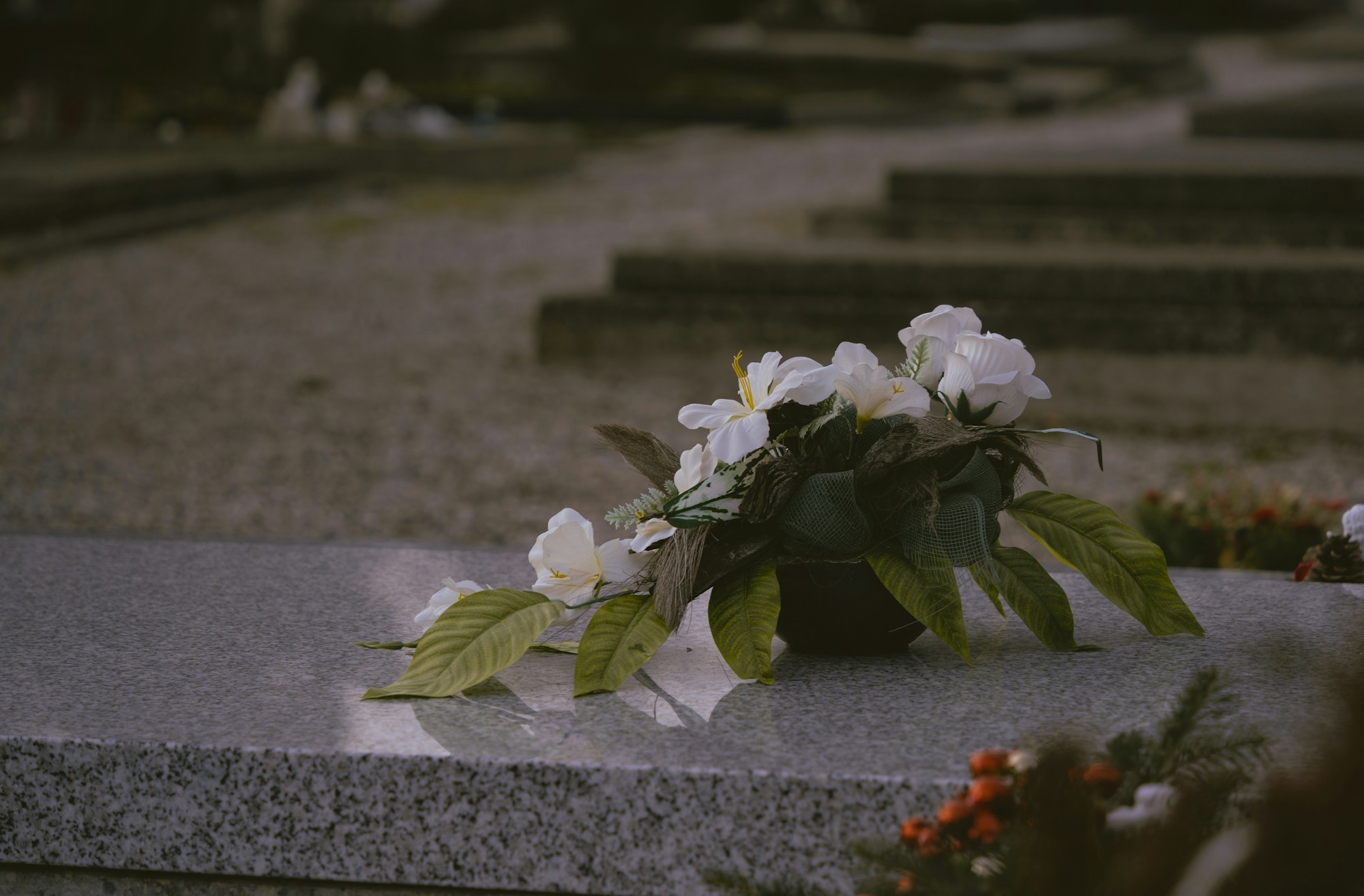 White lily flowers resting on a stone grave marker with mourners' legs and shoes visible in the background.