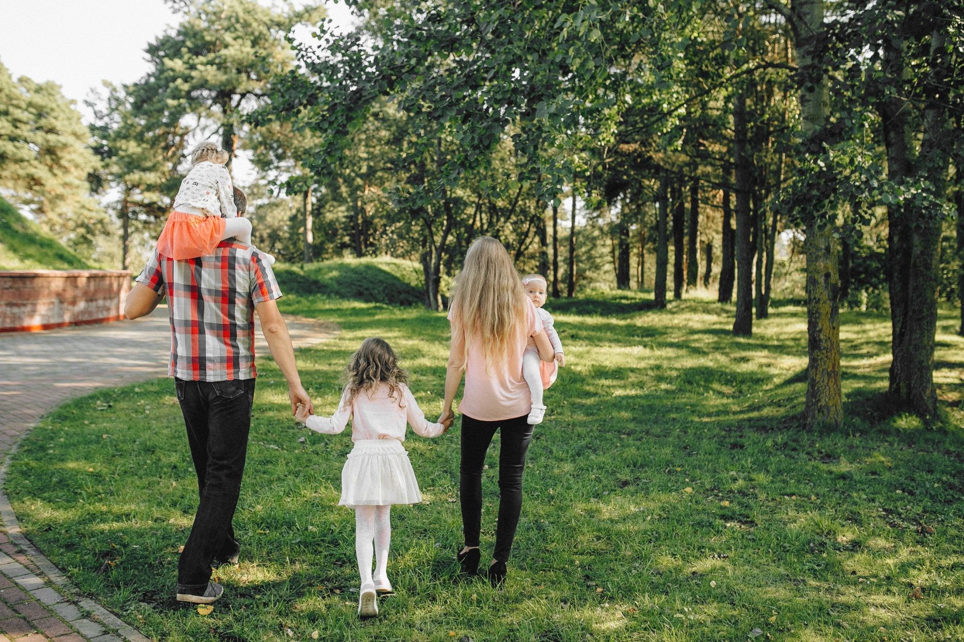 A family of five, seen from behind, walks hand in hand through a sunlit wooded park, two of the children being carried.
