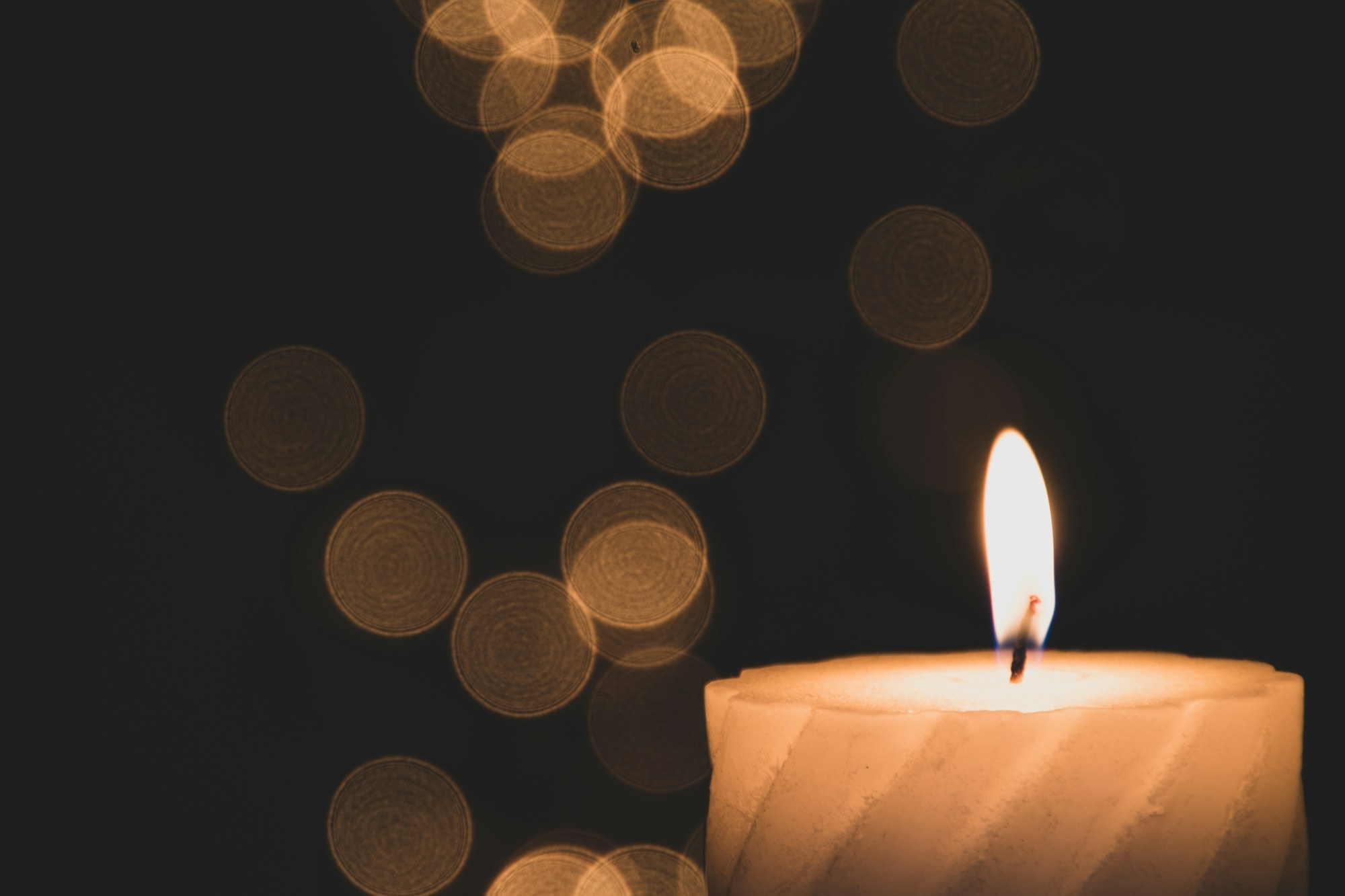 A single lit white candle glows in the foreground against a dark background with soft bokeh light.
