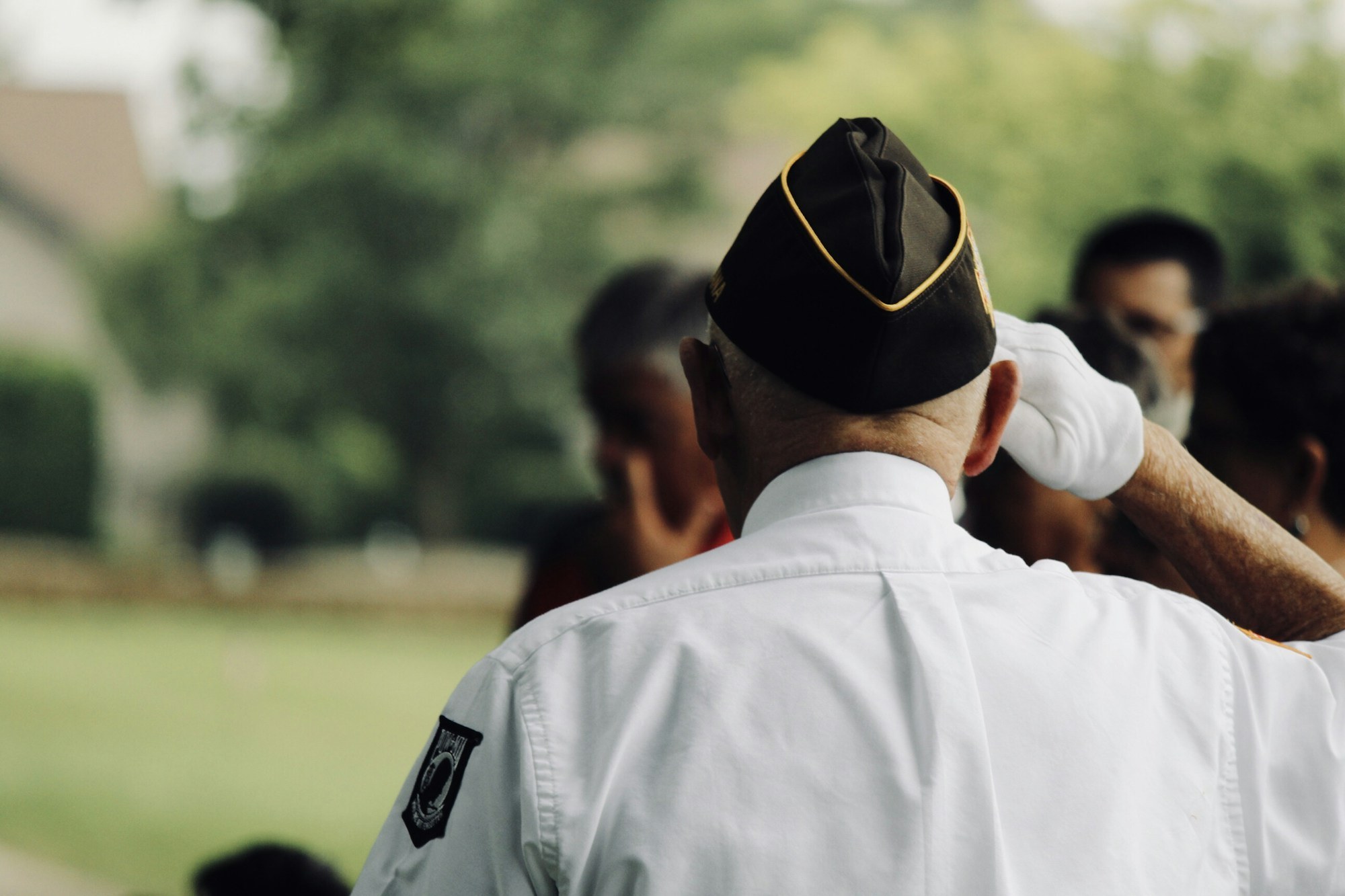 A man in a veteran's cap and white uniform shirt salutes during an outdoor ceremony, seen from behind.