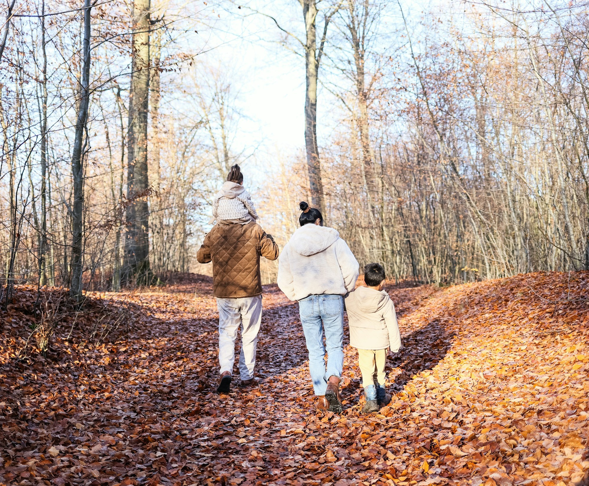 A woman and two young girls walking arm in arm along a sunny sidewalk near a stone wall.