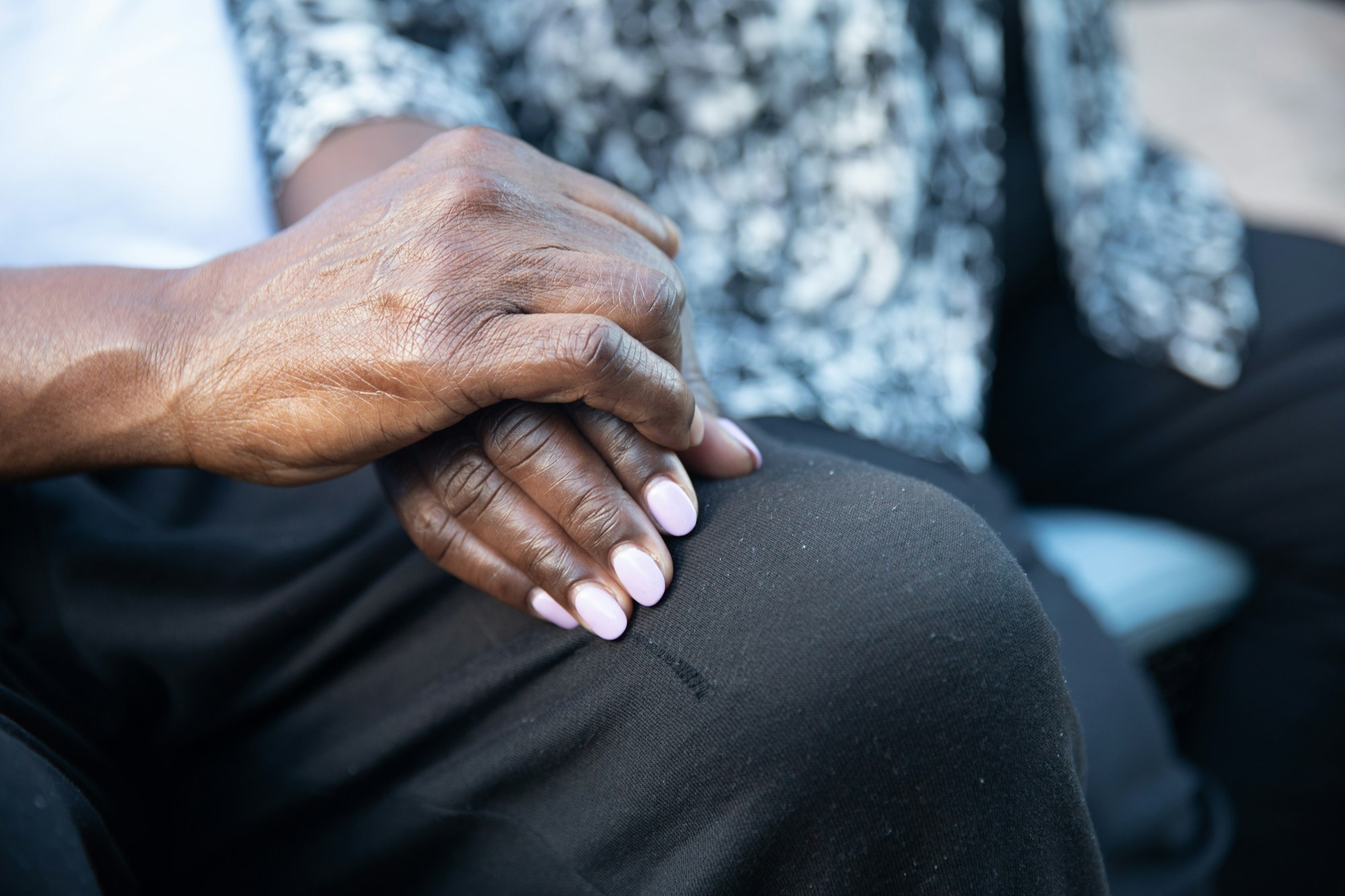 A close-up photograph of an elderly woman's hand resting calmly on her knee, dressed in dark clothing with a patterned cardigan.