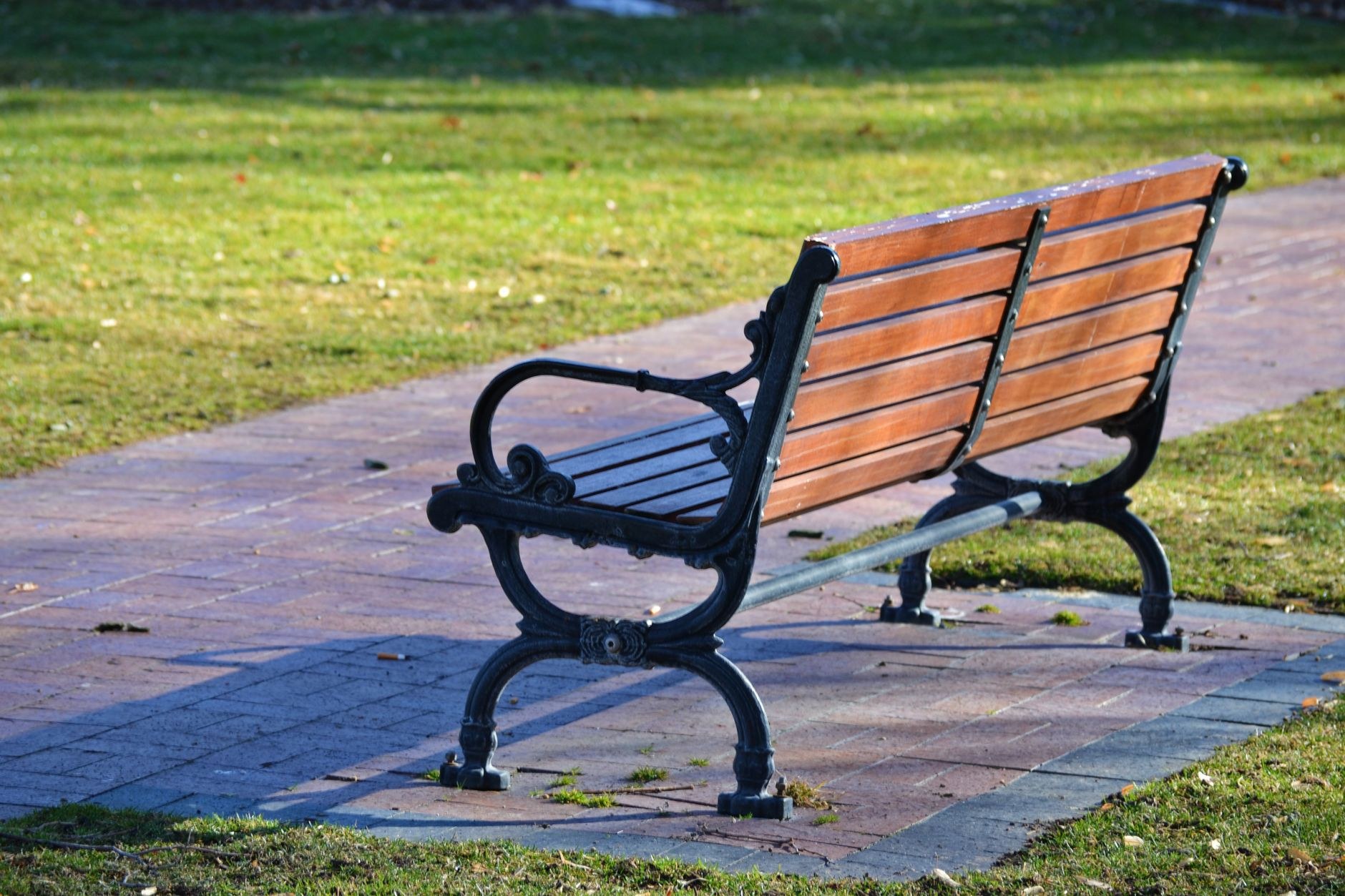 An empty ornate wooden and metal park bench sits on a brick path beside a grassy lawn.