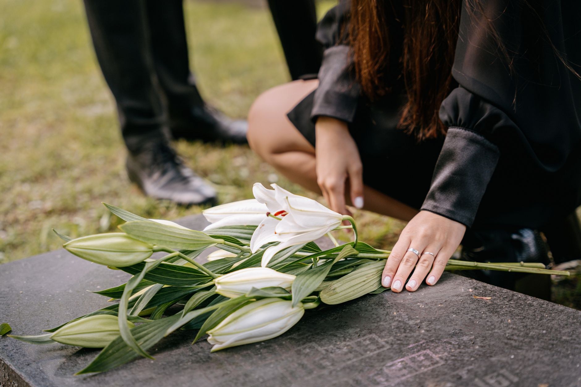 A woman's arm reaches down to place white lilies on a flat grave marker in a cemetery, with headstones and a large tree in the background.