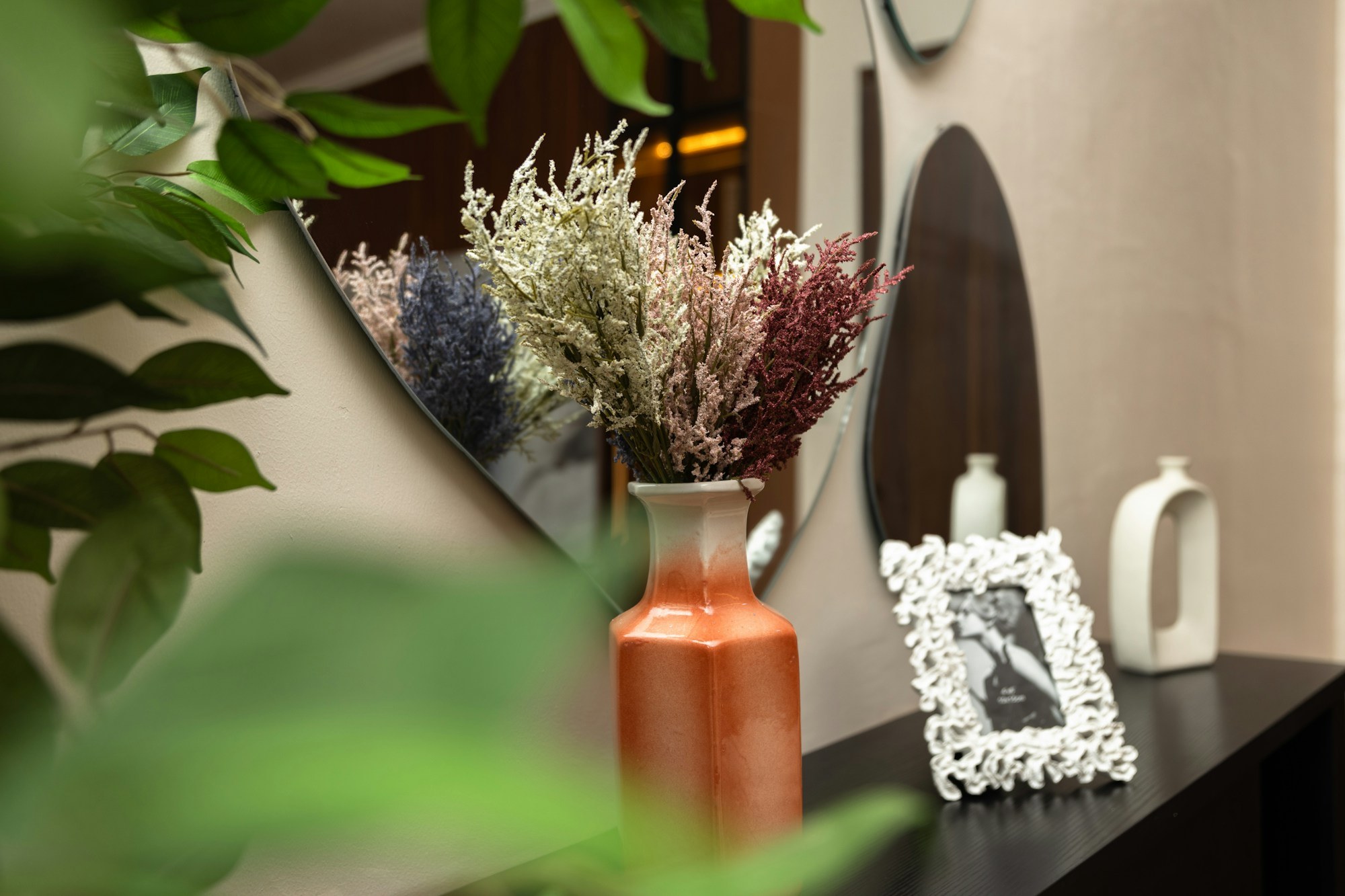 An orange ceramic vase with dried flowers sits on a dark console table beside a decorative picture frame, reflected in a curved wall mirror, with green plant leaves in the foreground.