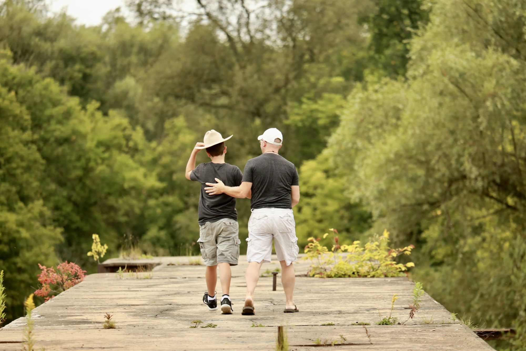 A man and teenage boy walk arm in arm together on a tree lined bridge, representing family bonds across generations.
