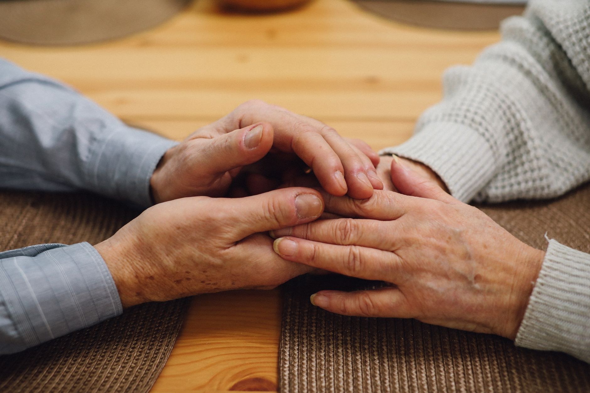 Close-up of two people's hands clasped together while seated on wooden chairs, one comforting the other.